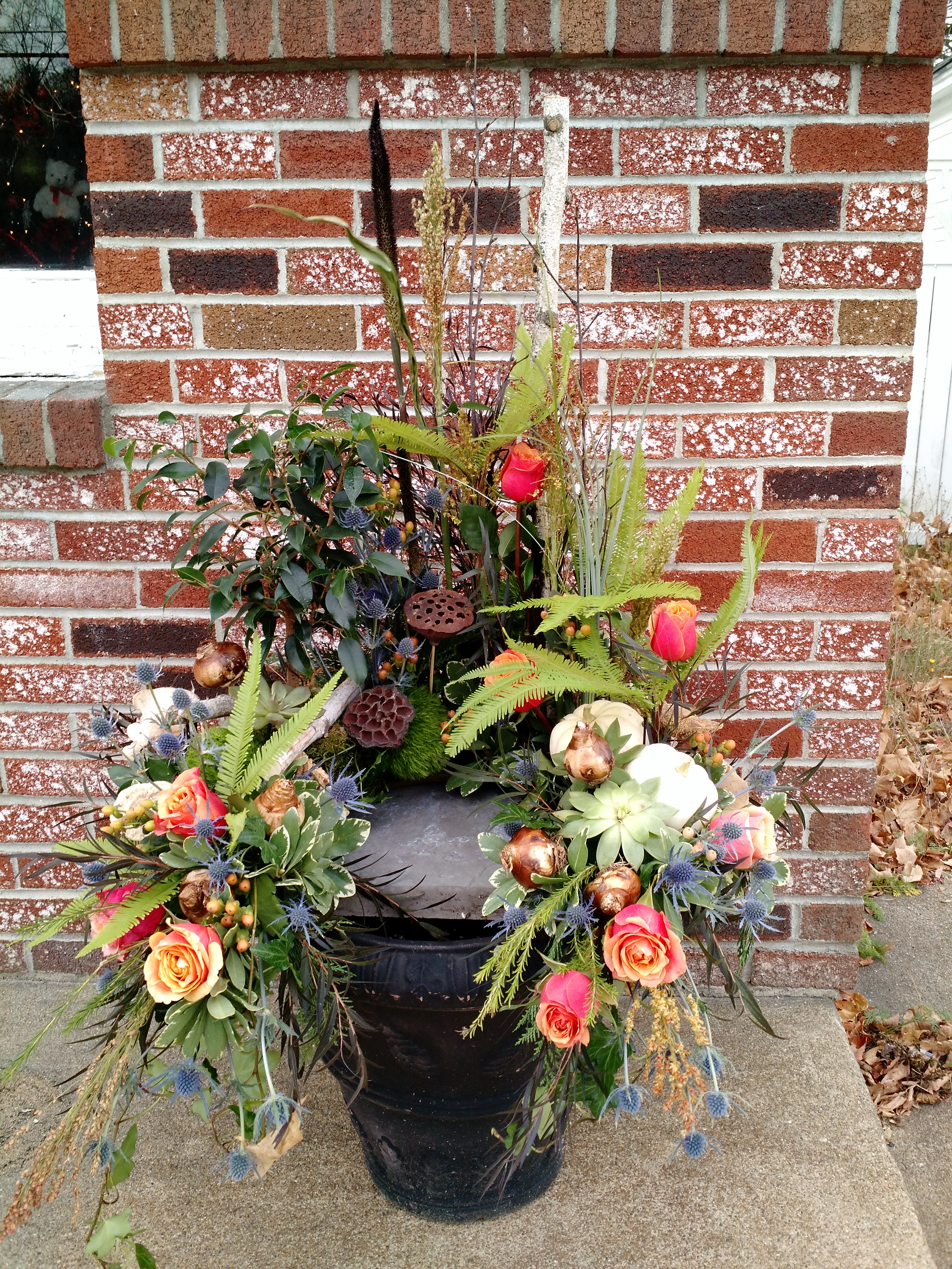 Outdoor arrangement of orange roses, succulents, and pods in a black urn against a brick wall.