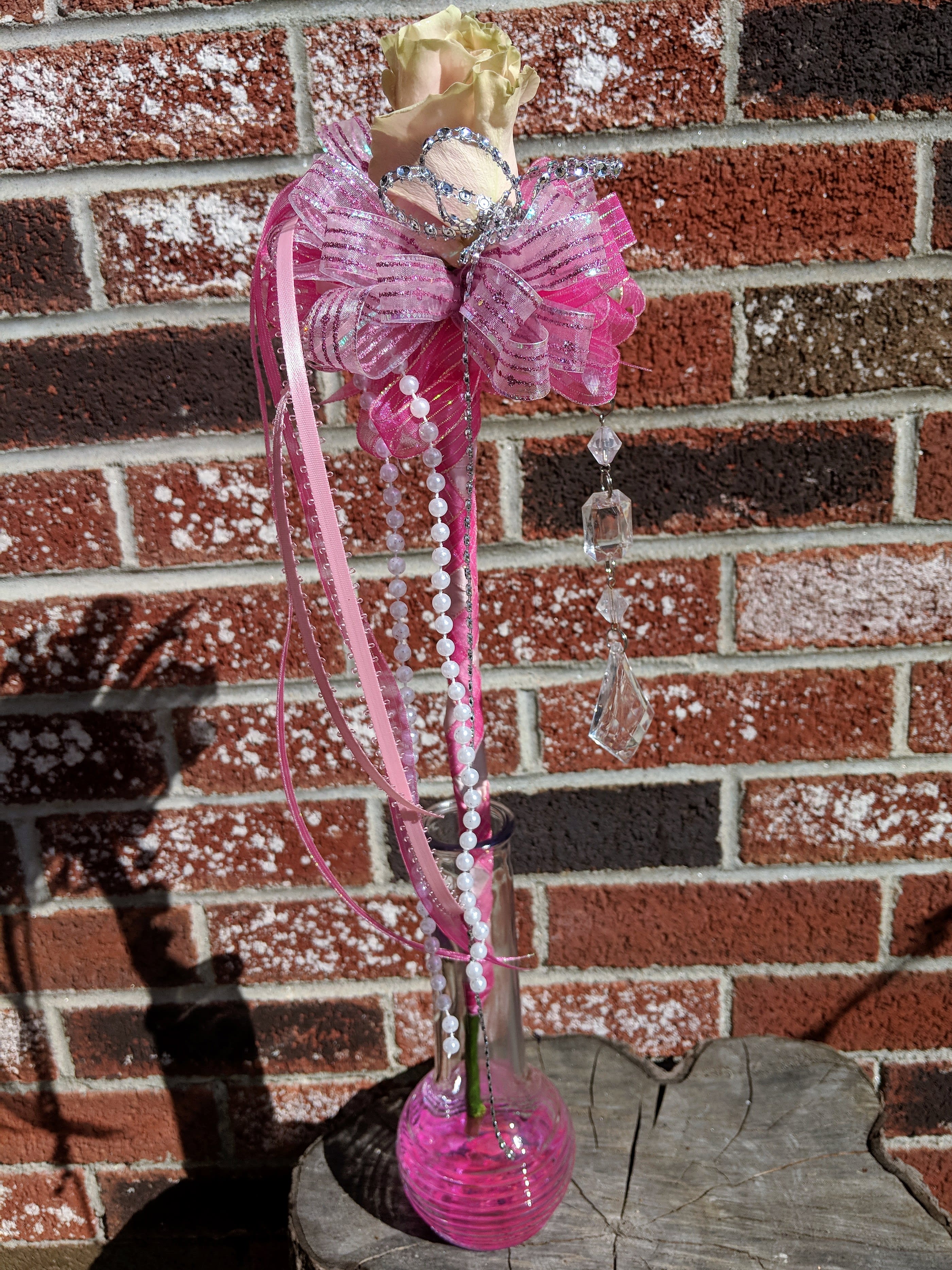 Single pale rose in a tall pink-accented glass bud vase with tiara, bow, pearls, and crystals.