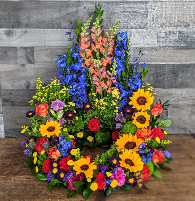 Tall circular wreath with bright sunflowers, roses, carnations, and blue delphinium.