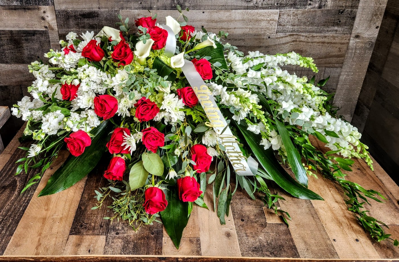 Large red and white casket spray with roses, lilies, and ribbon on a wooden surface