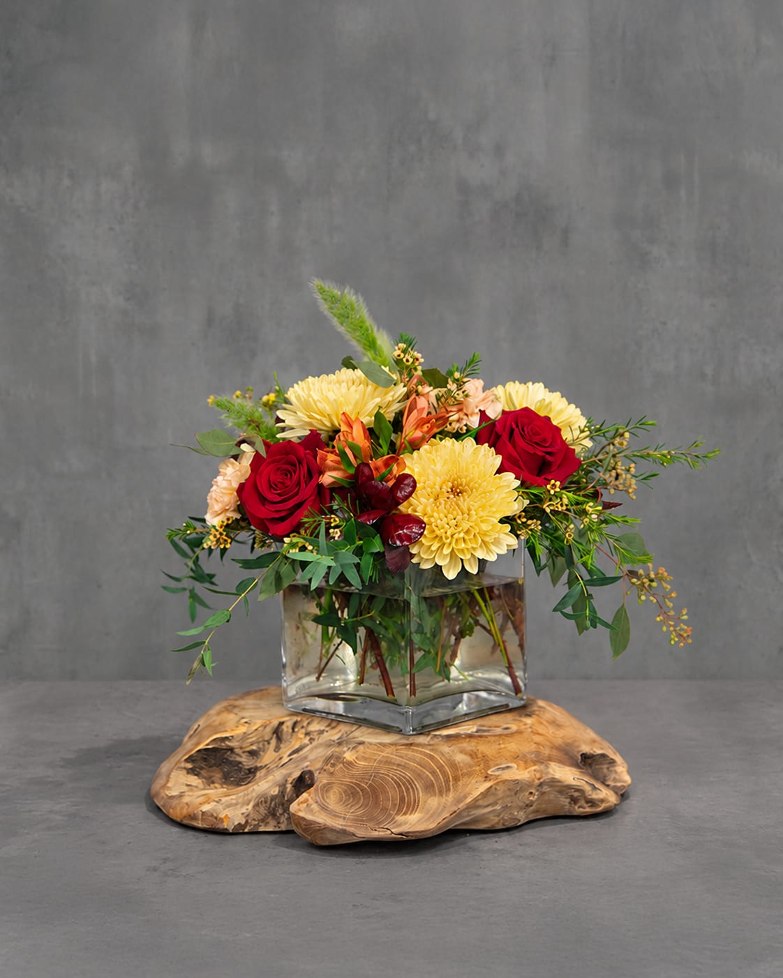 Red roses and yellow mums in a clear square vase on a wood slab