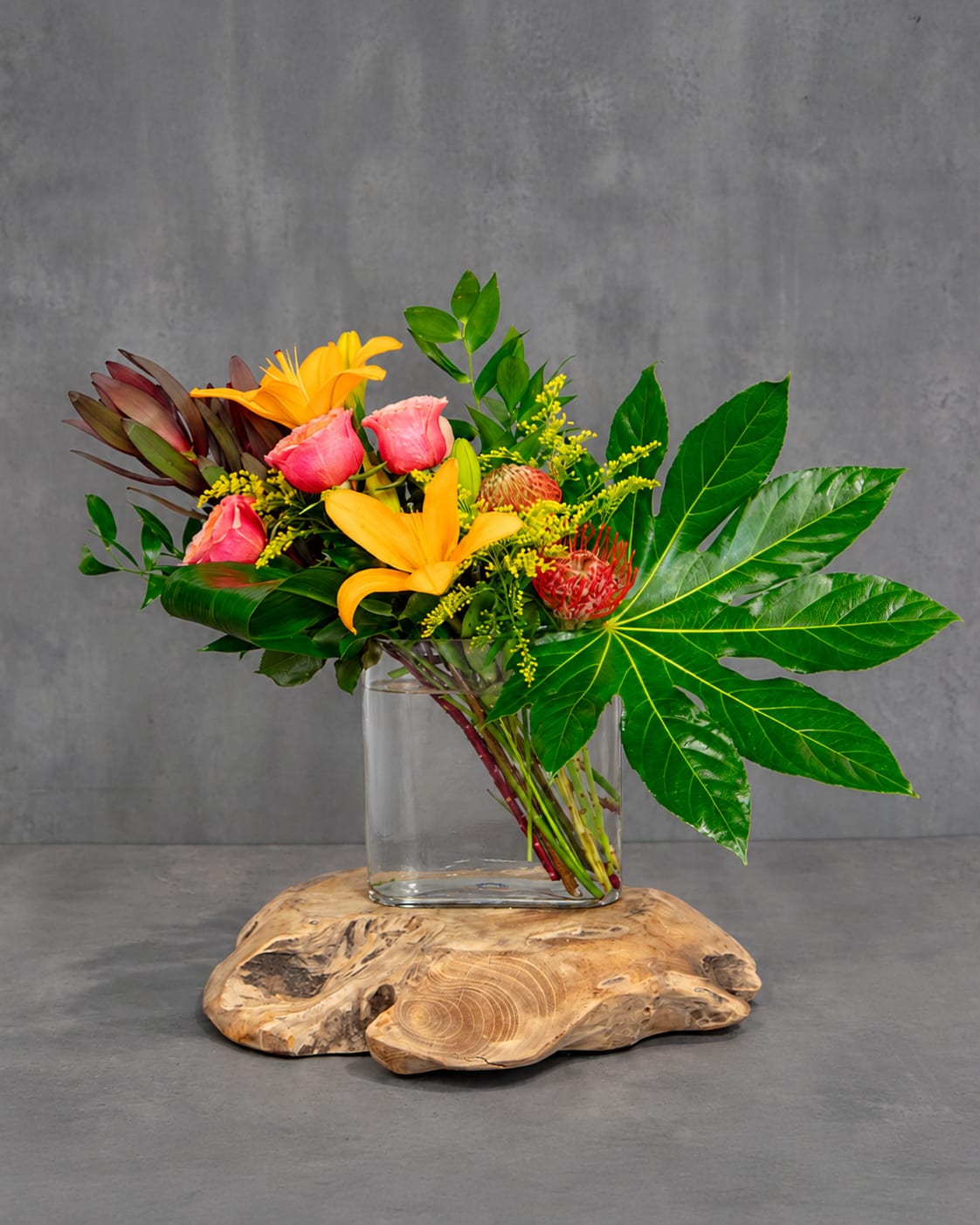 Tropical bouquet with yellow lilies, pink roses, and red protea in a glass vase