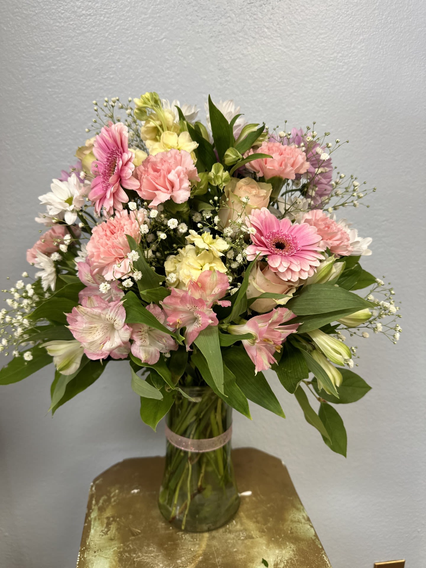 Pastel Dream Arrangement - A soft, airy cascade of pink gerbera daisies and creamy white carnations creates the kind of pastel statement that makes someone feel truly noticed. This flower arrangement is a one-sided bouquet designed for gifting - perfect as a birthday bouquet, a spring centerpiece, or a feminine celebration arrangement that reads cheerful and romantic from the room's primary viewpoint.  The arrangement pairs gerbera daisies, carnations, alstroemeria, daisies and delicate white filler blooms in a clear glass vase tied with a pink ribbon, blending bright pink accents with creamy whites, pale yellows and fresh greens for layered texture and a garden-fresh feel. Sized to sit on a table or desk, the rounded, one-sided composition directs attention toward the recipient while the clear vase keeps the look light and modern. Care: keep fresh water in the vase, trim stems every few days, and avoid direct heat to extend bloom life.