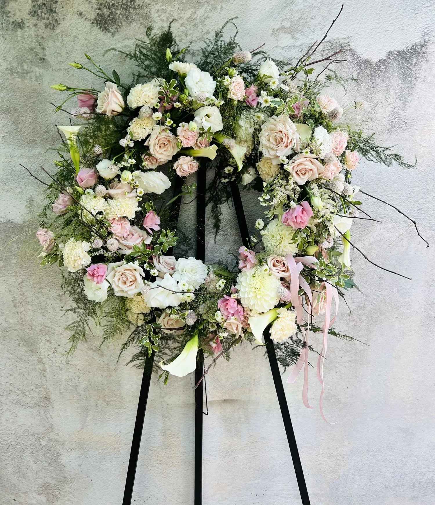 Garden Memorial Wreath - Honor the memory of a loved one with our white and pink garden-style memorial wreath, thoughtfully designed on a classic easel. This elegant tribute features premium seasonal florals and lush greenery, arranged in a soft, natural style that evokes peace and remembrance. Each wreath is handcrafted with care, blending fresh blooms in serene whites and gentle pinks to create a heartfelt expression of sympathy. Perfect for memorial services, celebrations of life, or graveside tributes, this timeless design offers a beautiful way to share your love and condolences.  Please Note  Deluxe option pictured