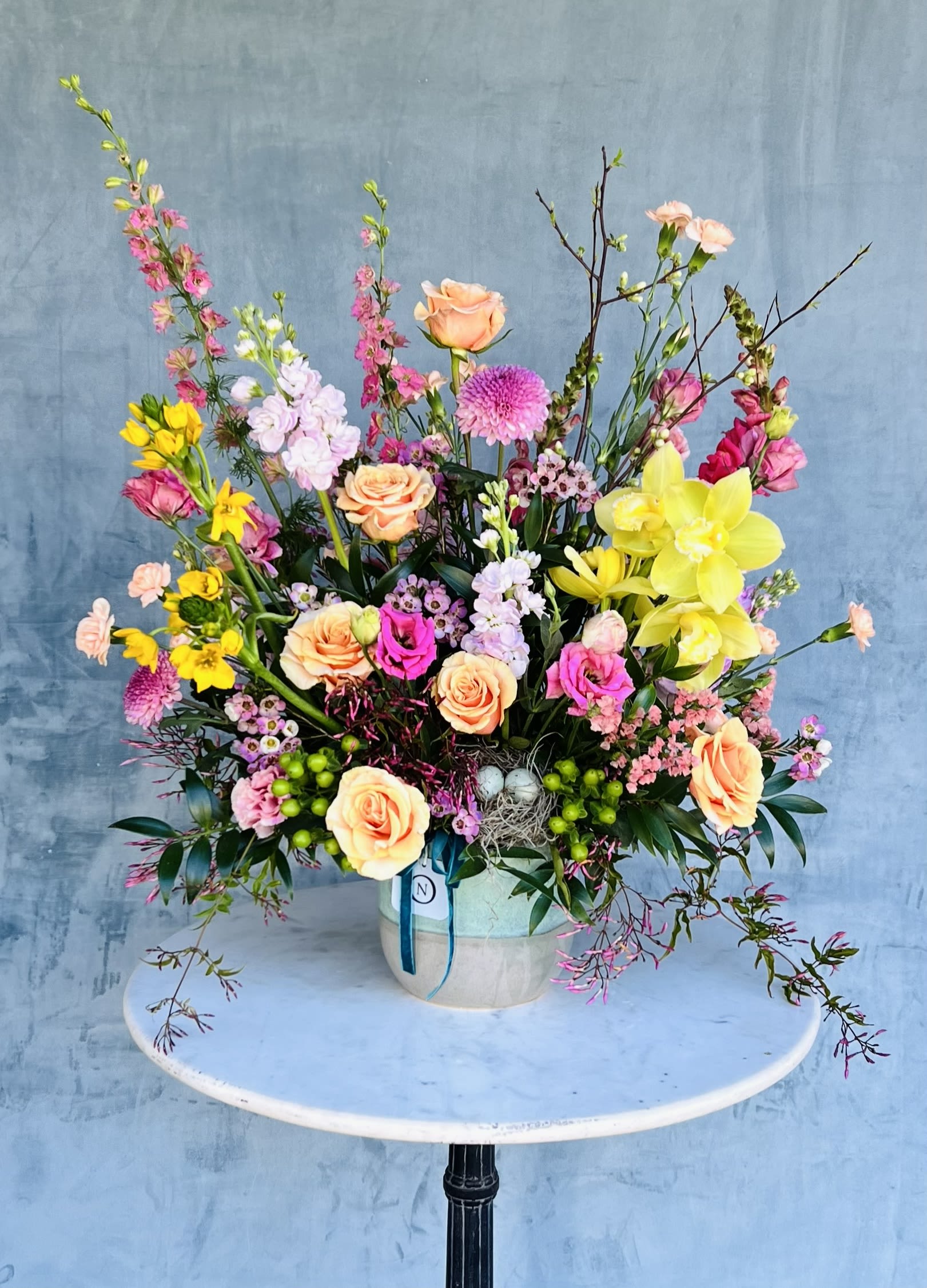 Rainy - This garden-style spring arrangement is designed to feel bright, cheerful, and full of natural movement. Featuring premium seasonal florals in vibrant shades of pink, yellow, and peach, each bouquet is carefully handcrafted with roses and orchids as standout focal blooms.  The design embraces an airy, garden-inspired style with lush greenery woven throughout, creating texture, depth, and a freshly gathered feel. Every arrangement is made to order using the finest seasonal flowers available, ensuring each piece feels unique and full of life.  Flowers and greenery may vary due to seasonal availability, but that’s part of the charm! Our creative designers will substitute with the freshest high end product available- some grown in our very own garden!- and create something beautiful.  *Have a special request?* Let us know in the special instructions section during checkout.