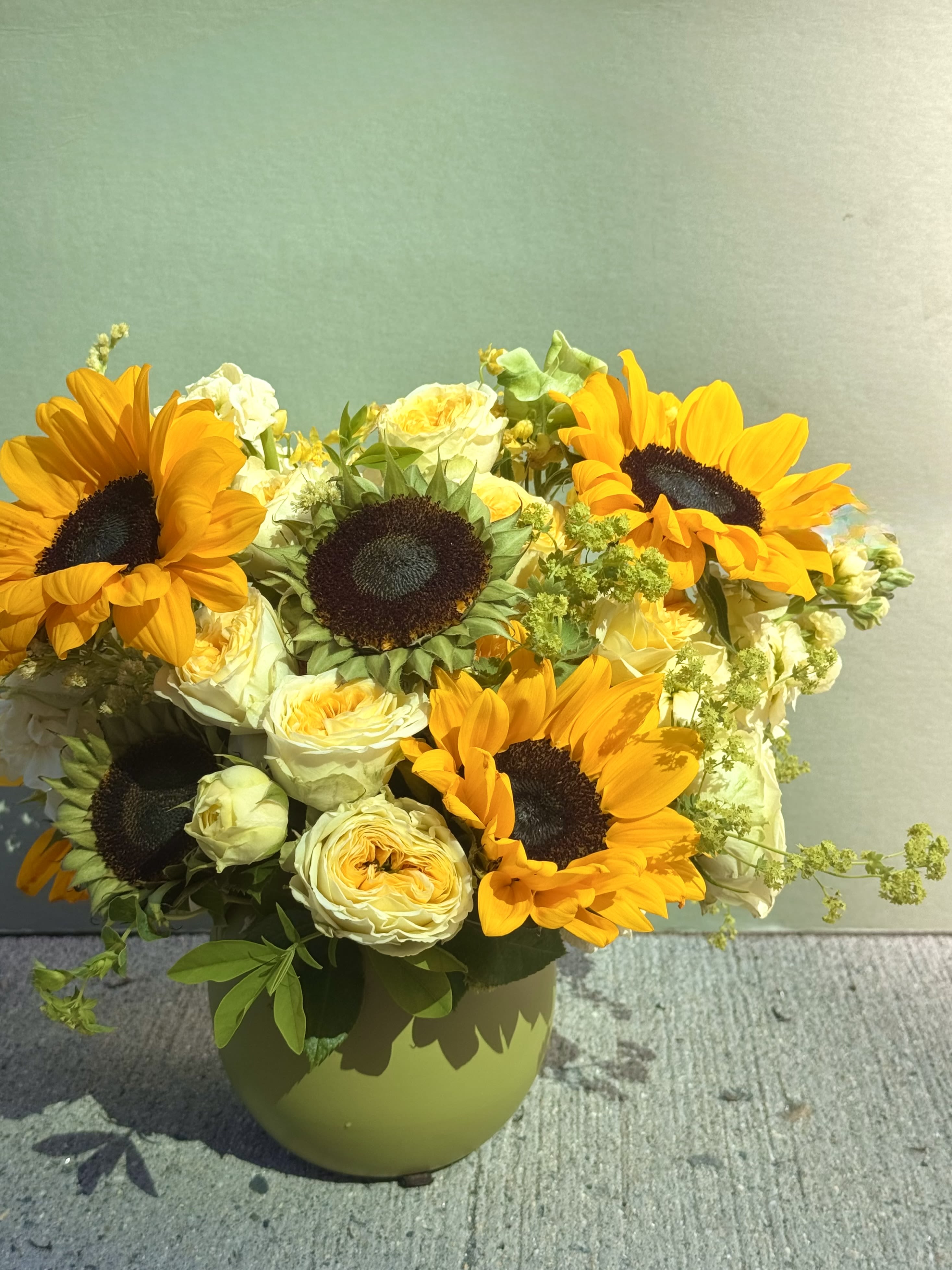 Sunflowers season - Golden sunflowers take center stage in this sunflower arrangement for birthdays, thank-yous, or bright everyday gifting. The large, warm faces of the sunflowers paired with creamy white roses create an uplifting contrast that reads cheerful and sincere-the kind of bouquet that makes the person you're thinking of feel seen and celebrated.  Tucked among small yellow filler blooms and fresh greenery, the flowers are arranged in a rounded, full composition that feels abundant without being fussy. The stems come set in a matte green ceramic vase that's both simple and sturdy, about a medium tabletop size, perfect for desks, kitchen tables, or an entryway. Colors: sunny yellow, soft white, and garden green; flowers: sunflowers, roses, and seasonal fillers.  Ideal for brightening someone's day, saying thanks, or marking a casual celebration, this arrangement arrives ready to display. To keep it fresh, refresh the water every other day and trim stems slightly when you change the water. Order with same-day delivery when available for an easy, joyful surprise.