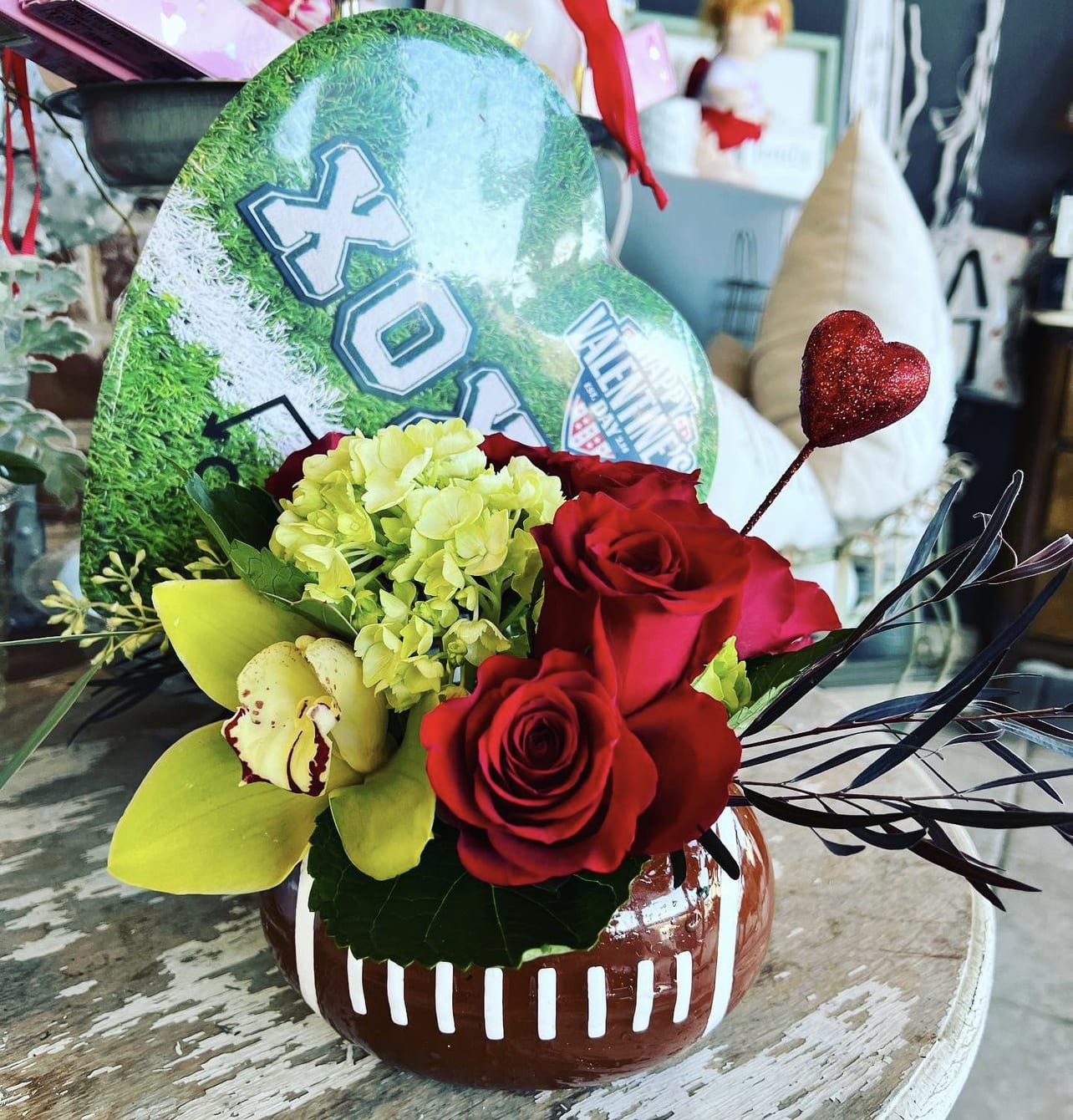 Red roses and yellow orchids in a football-shaped vase with a glitter heart pick