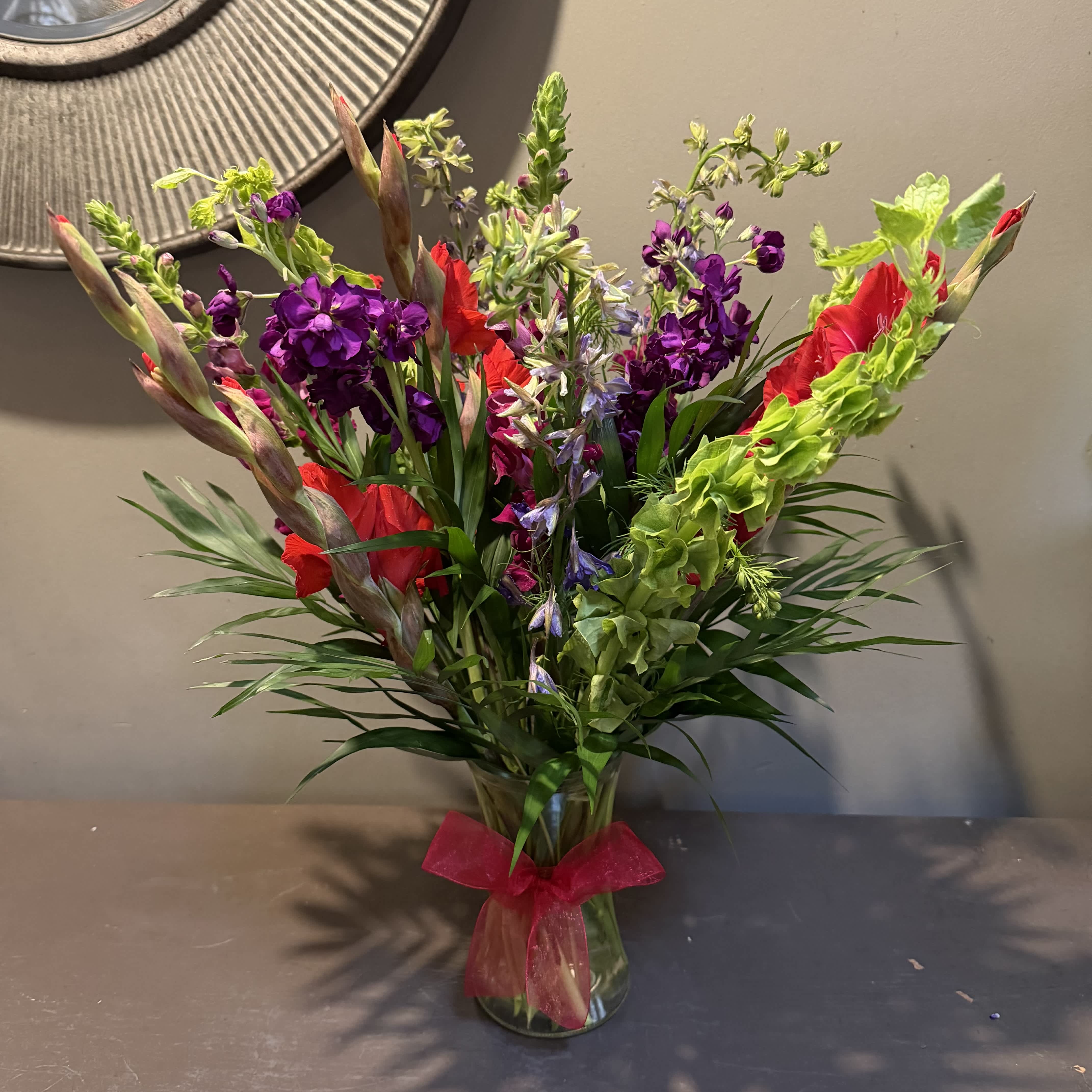 Tall mixed bouquet of red, purple, and green flowers in a glass vase with a pink ribbon.