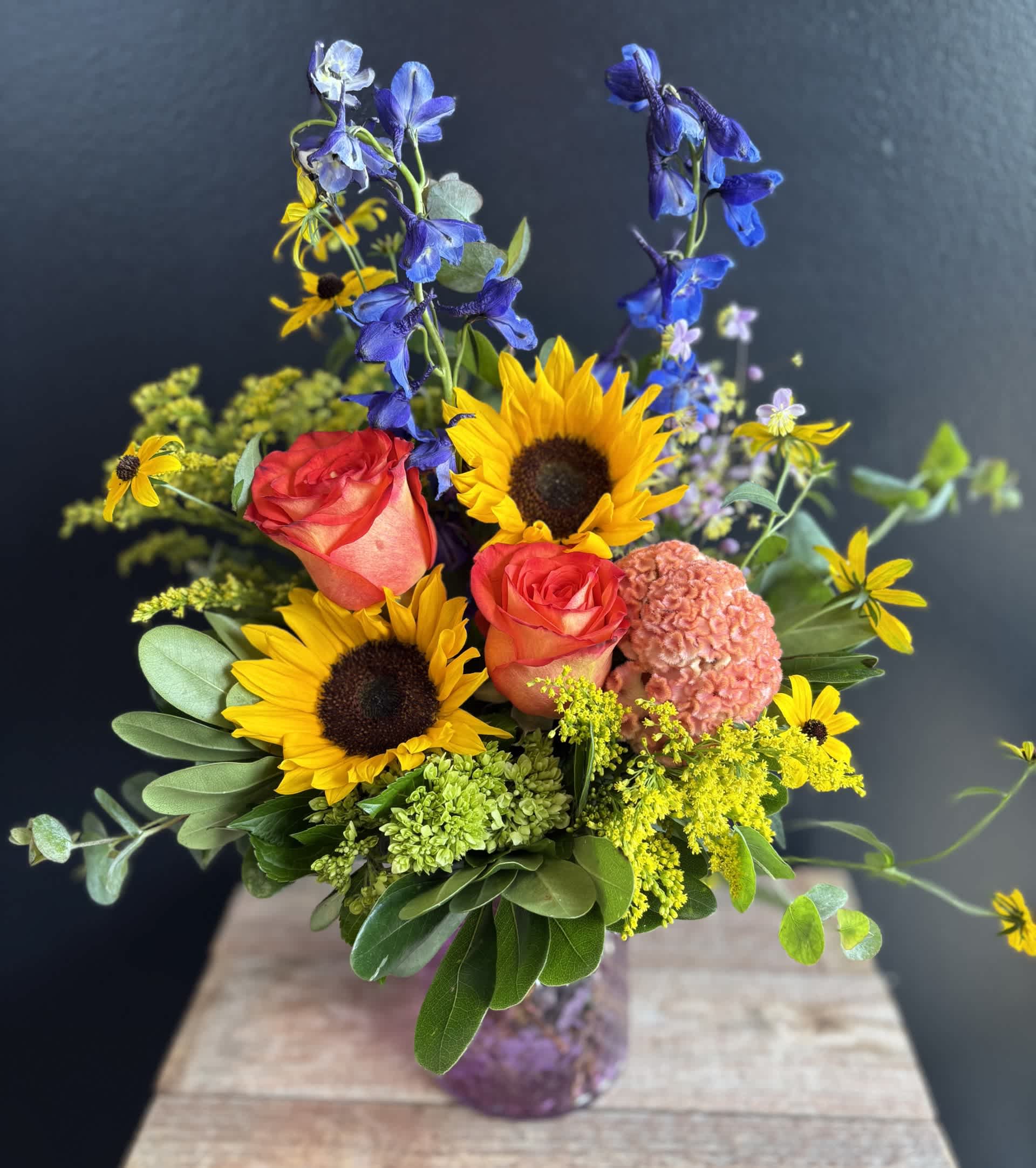 Bouquet with sunflowers, coral roses, and blue delphinium in a glass vase