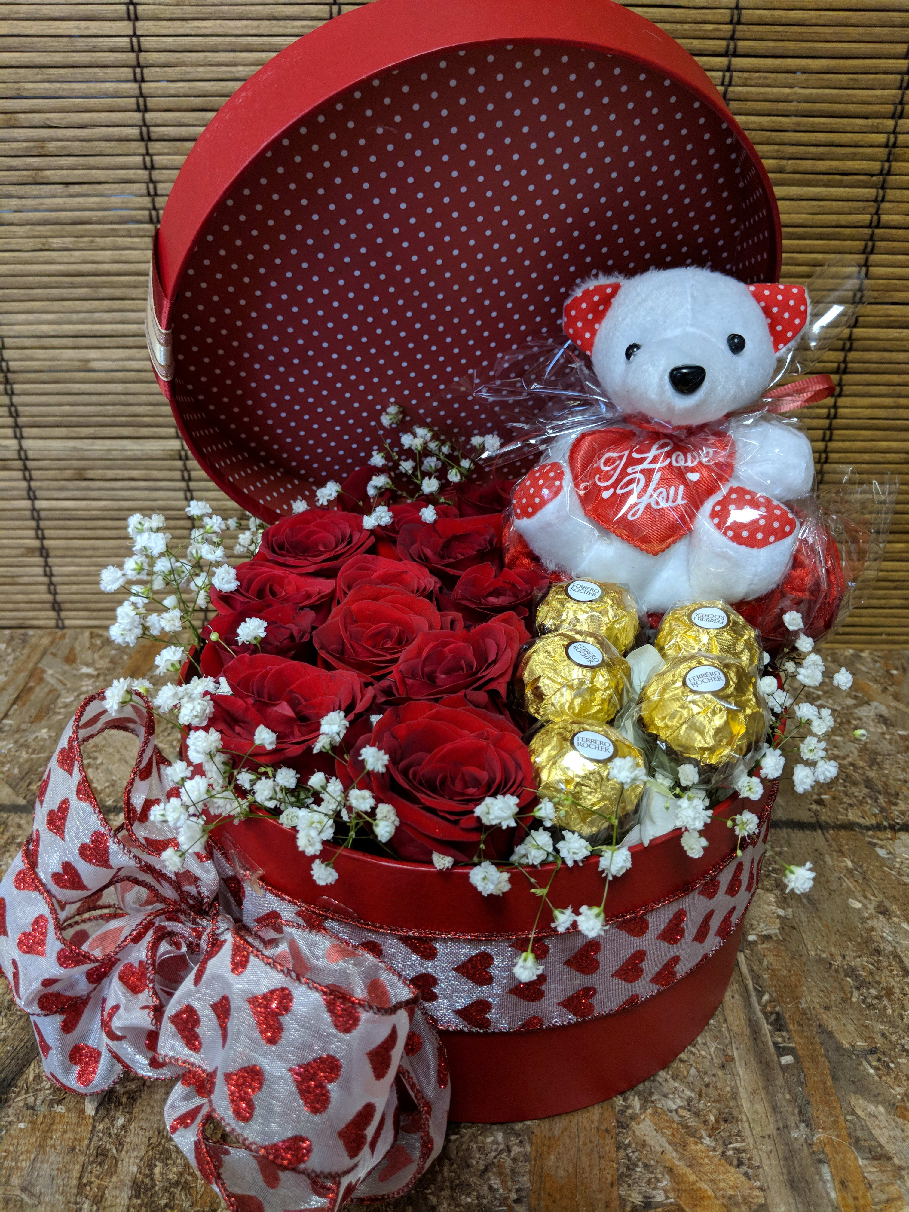Red hat box filled with red roses, gold-wrapped chocolates, and a white teddy bear holding a heart.