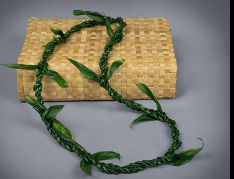 Braided green leaf lei draped over a tan woven mat against a gray background