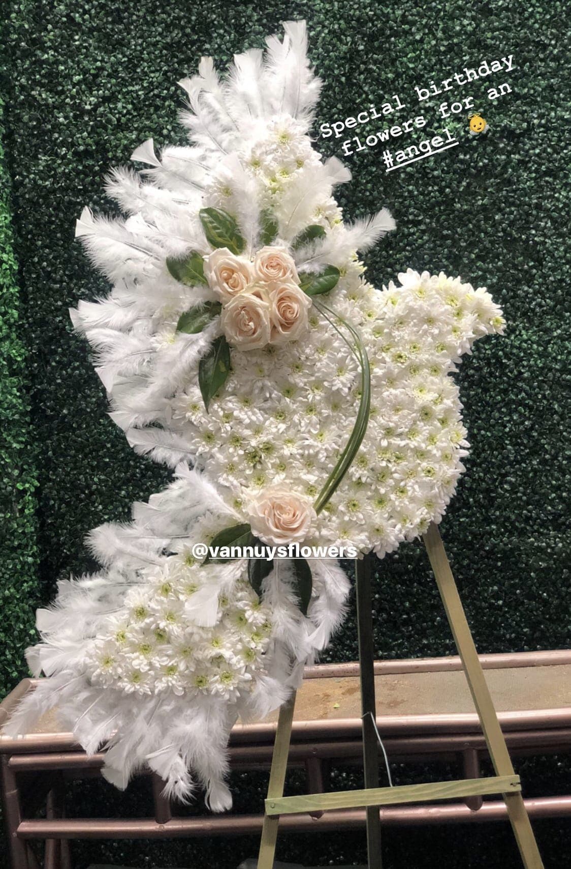 Angel-shaped standing spray of white flowers with blush roses and feathers on a metal easel