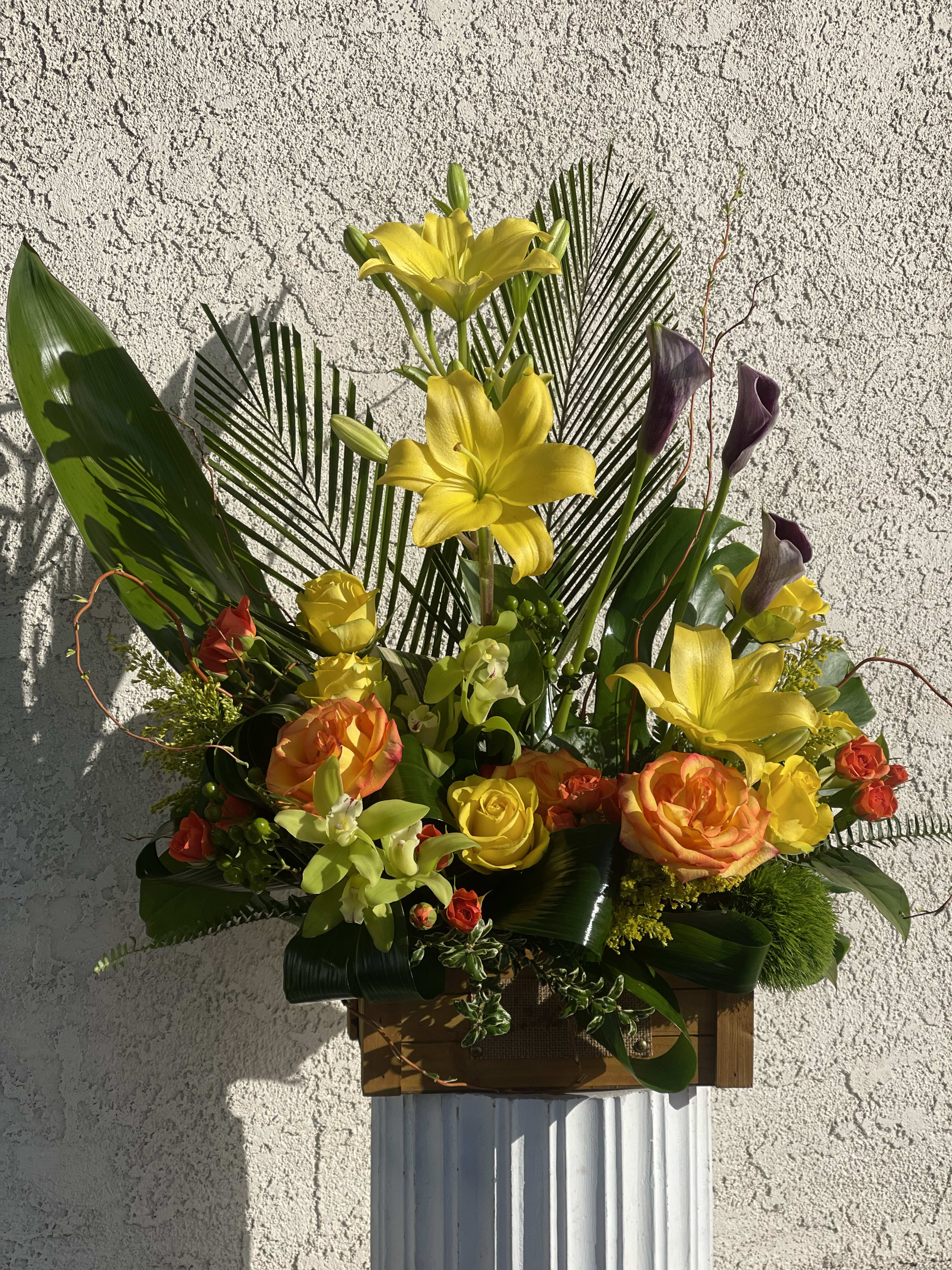 Tall arrangement of yellow lilies, orange and yellow roses, and green orchids in a wooden box