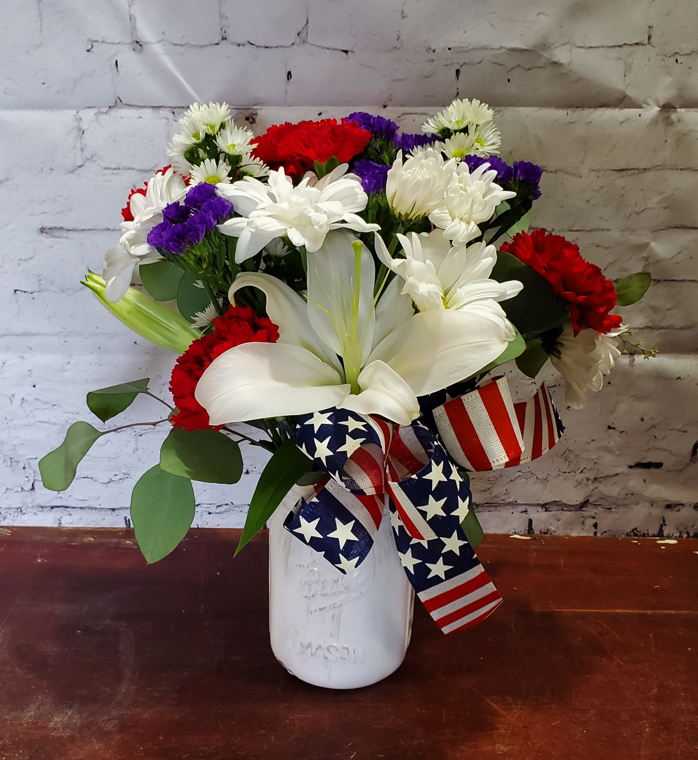 Bouquet of white lilies, red carnations, and purple daisies in a white vase with patriotic ribbon