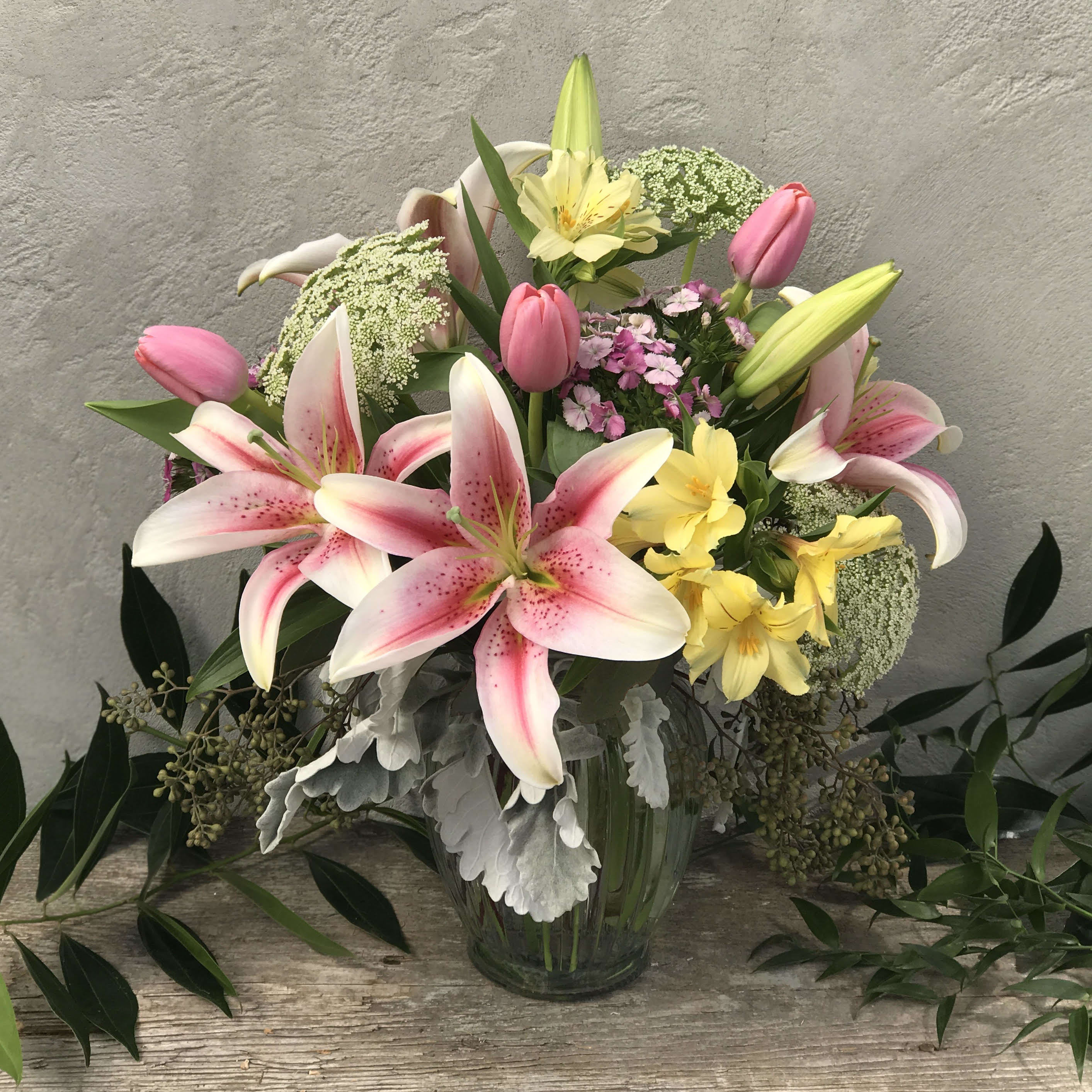 Pink lilies, tulips, and yellow flowers arranged in a clear glass vase against a neutral background