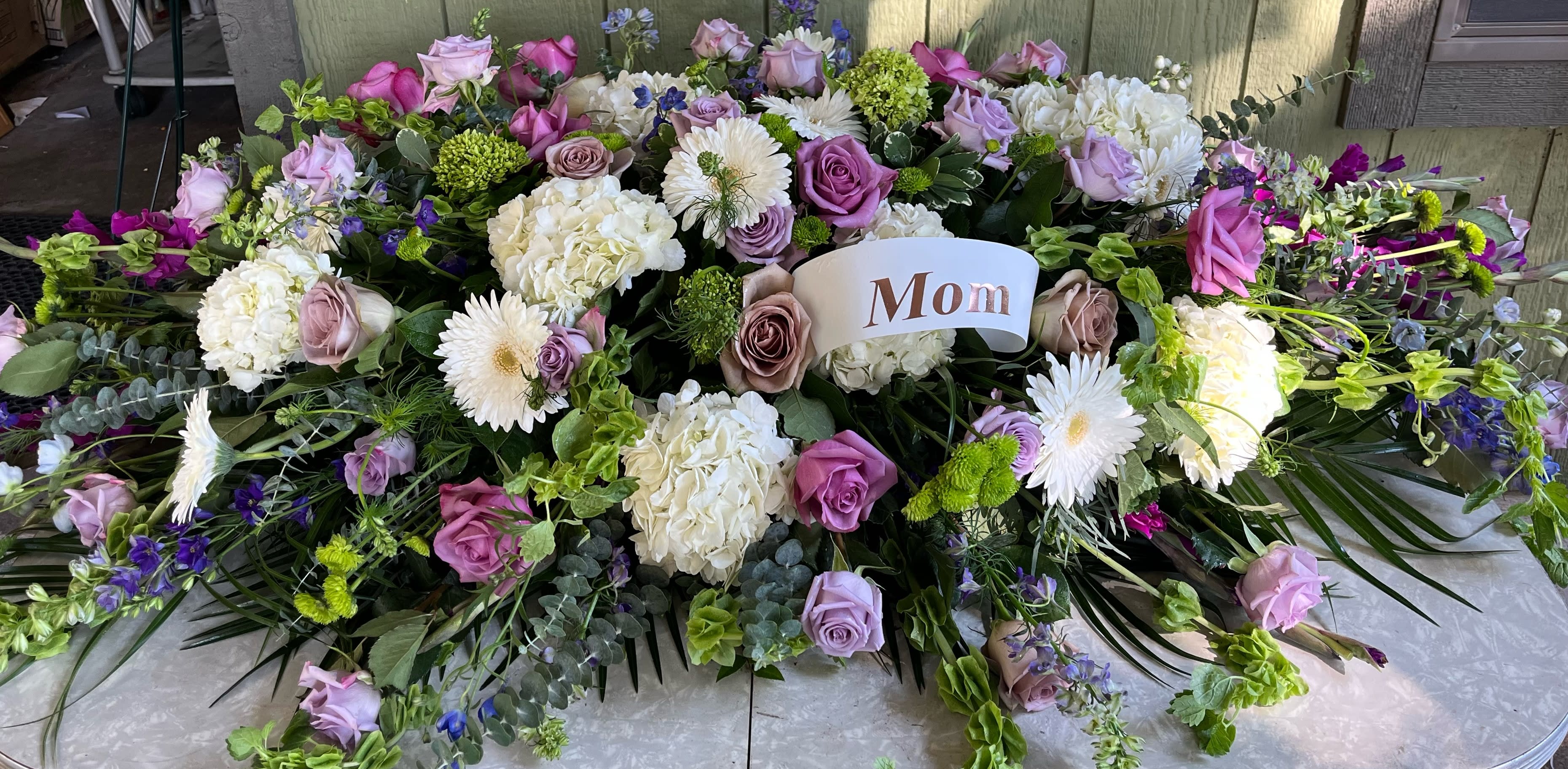 Large floral tribute with pink, lavender, white, and green blooms and a "Mom" card