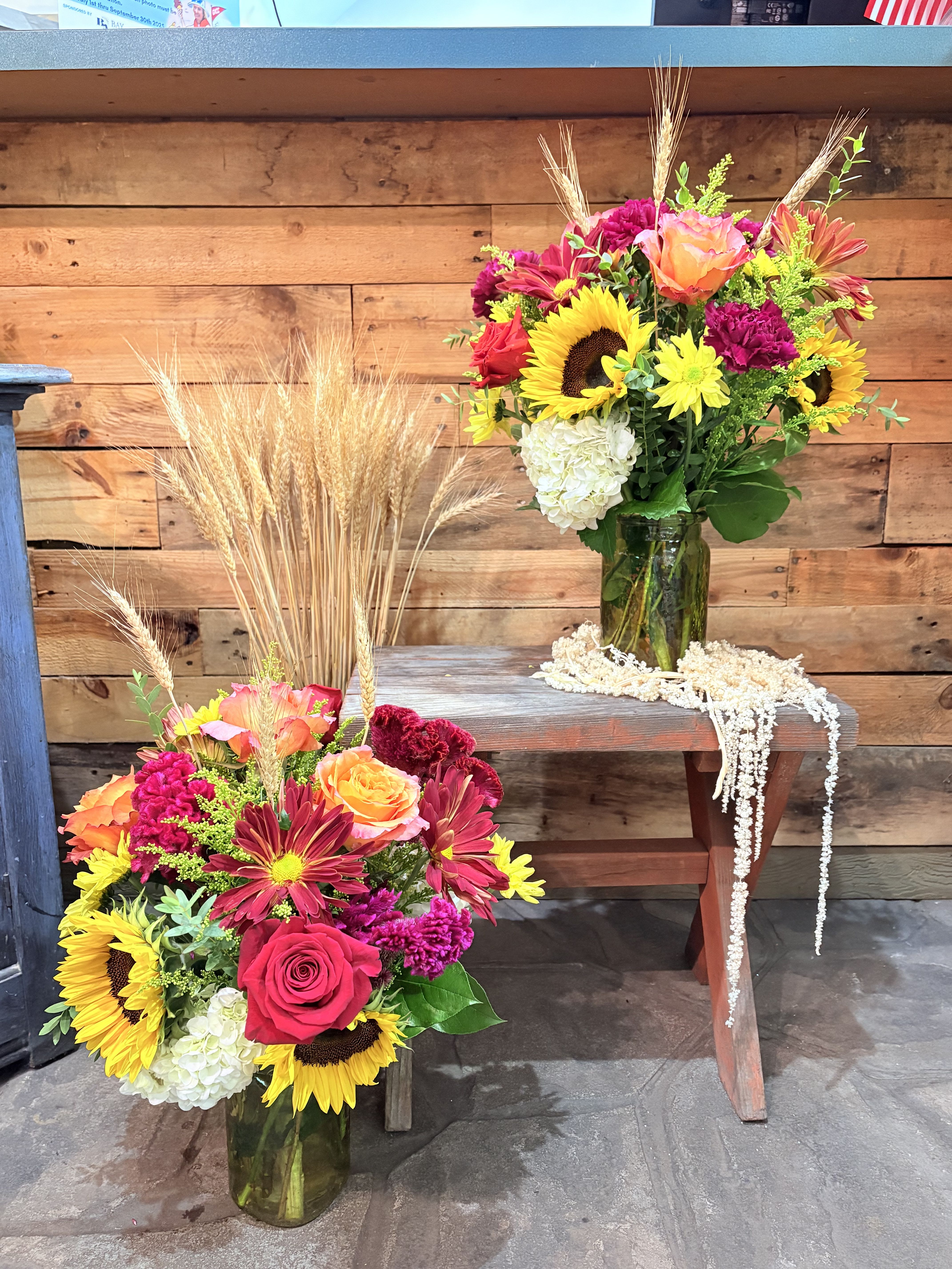 Two colorful flower arrangements in green glass vases with wheat accents