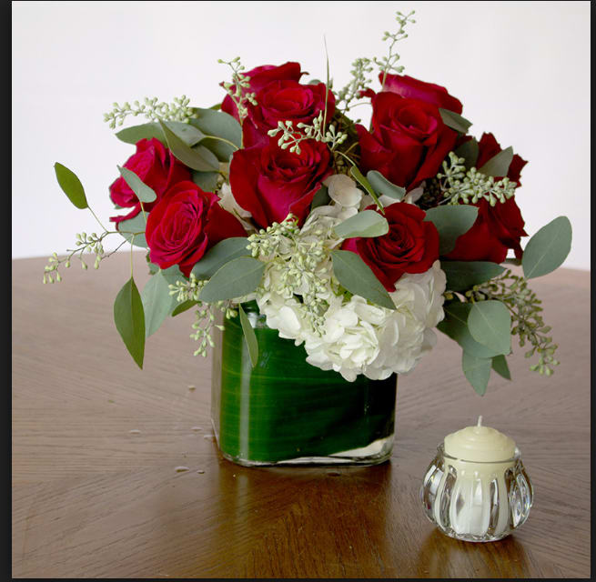 Red roses and white hydrangeas in a glass vase with a small candle beside it