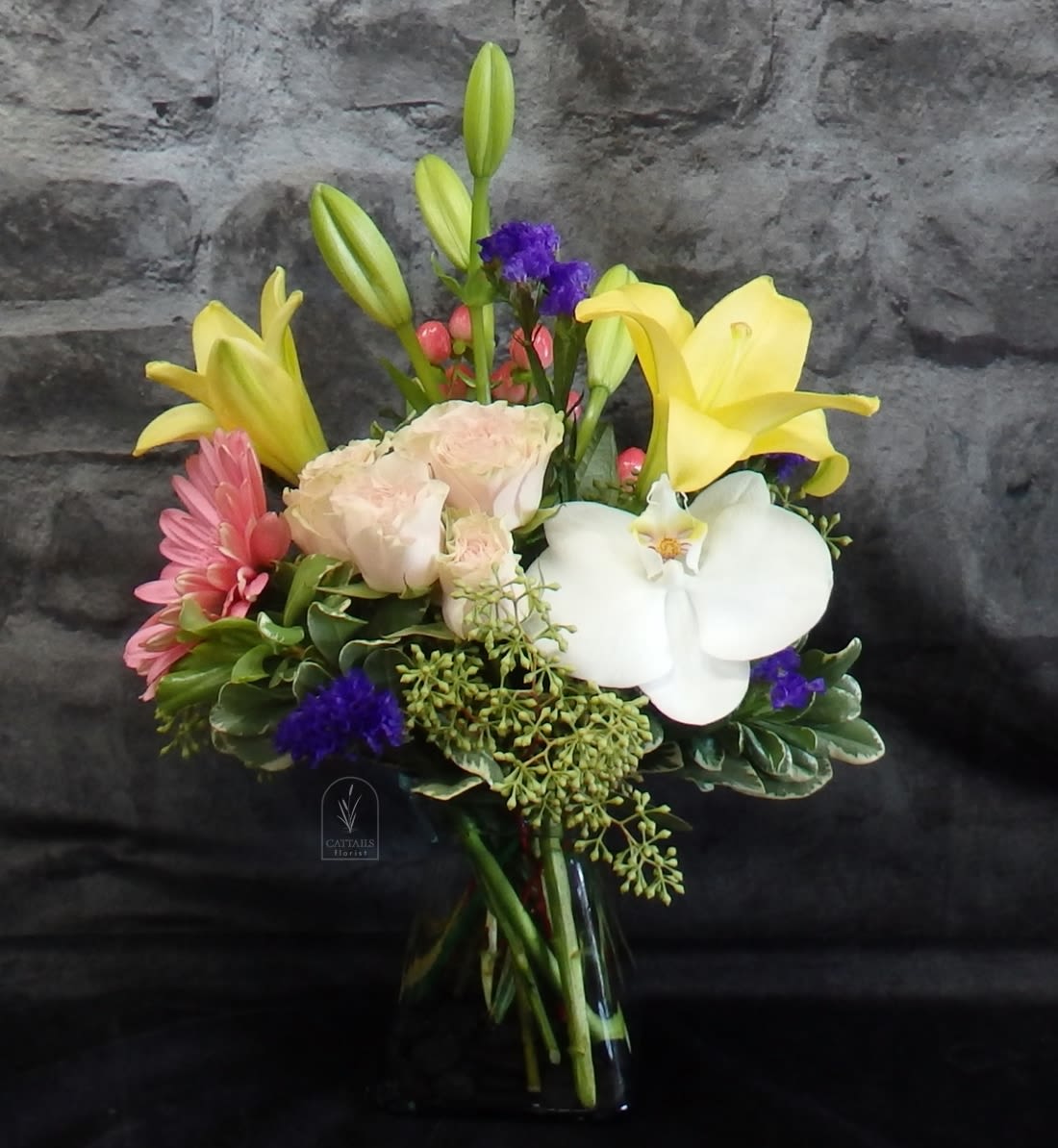 Mixed bouquet with yellow lilies, white orchid, pink blooms, and pale roses in a glass vase