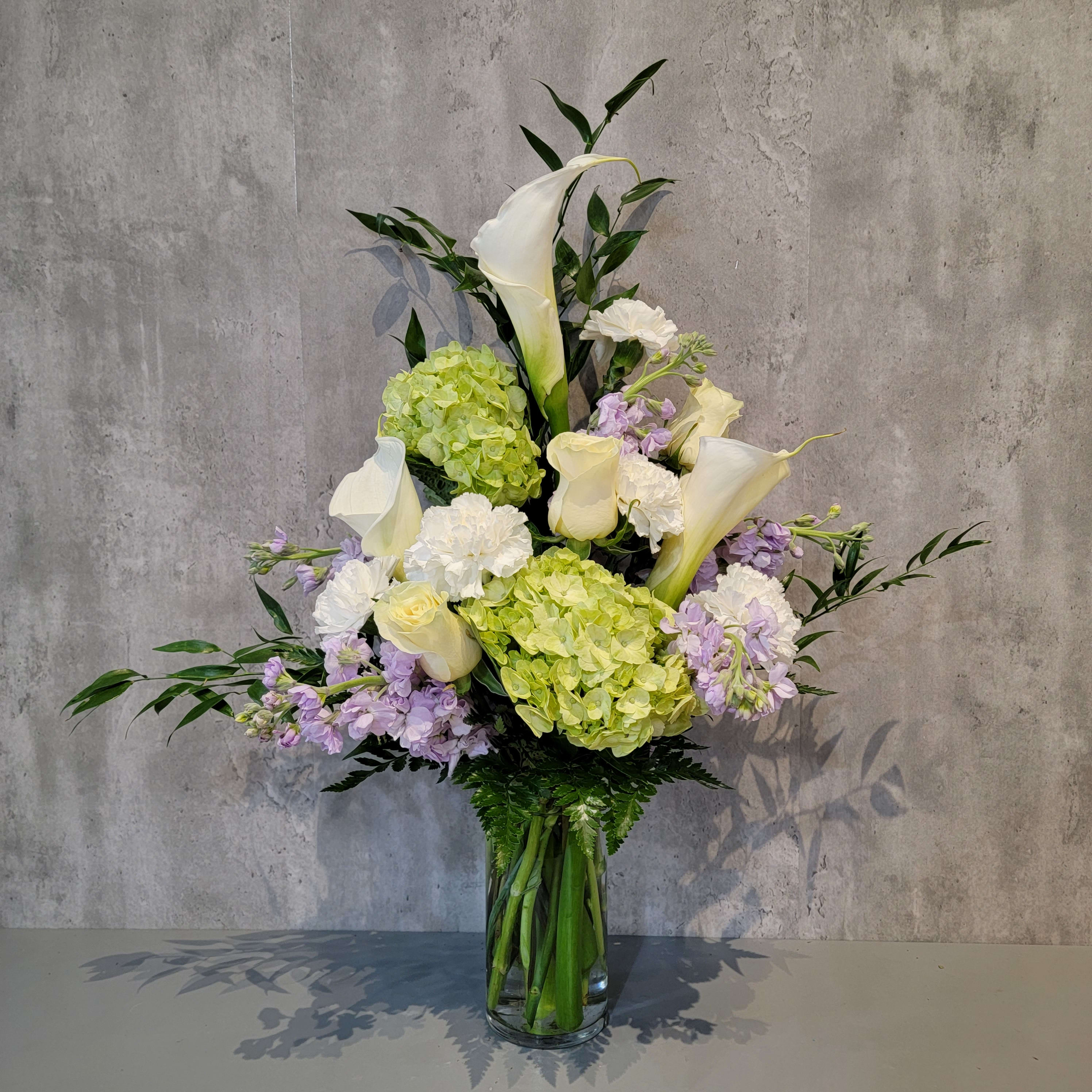 Tall bouquet of white calla lilies, hydrangeas, and lavender flowers in a glass vase