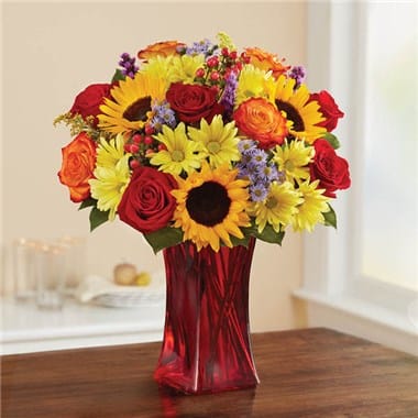 Mixed bouquet of sunflowers, roses, and daisies in a red glass vase