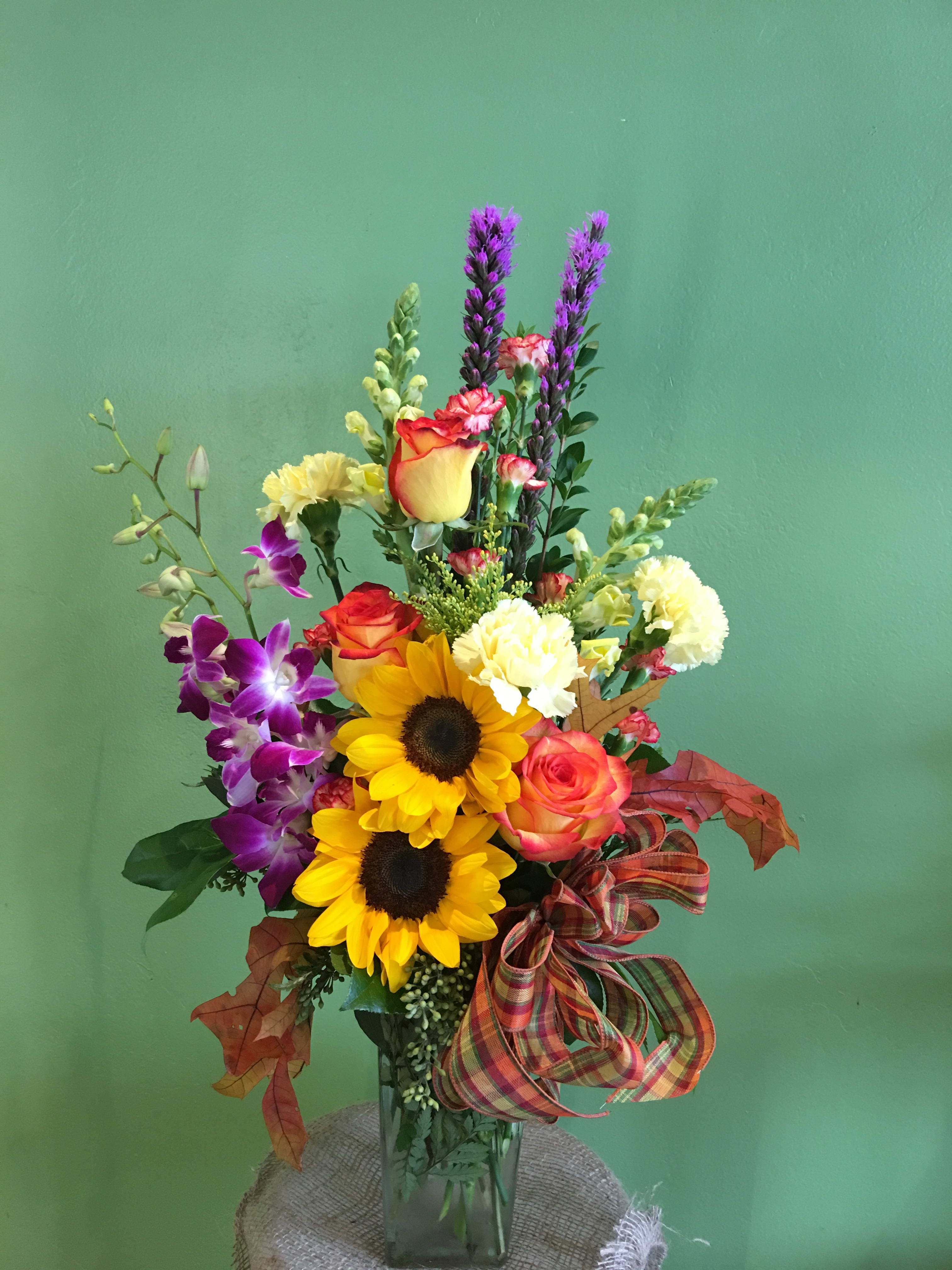 Mixed bouquet with sunflowers, roses, orchids, and purple spikes in a glass vase