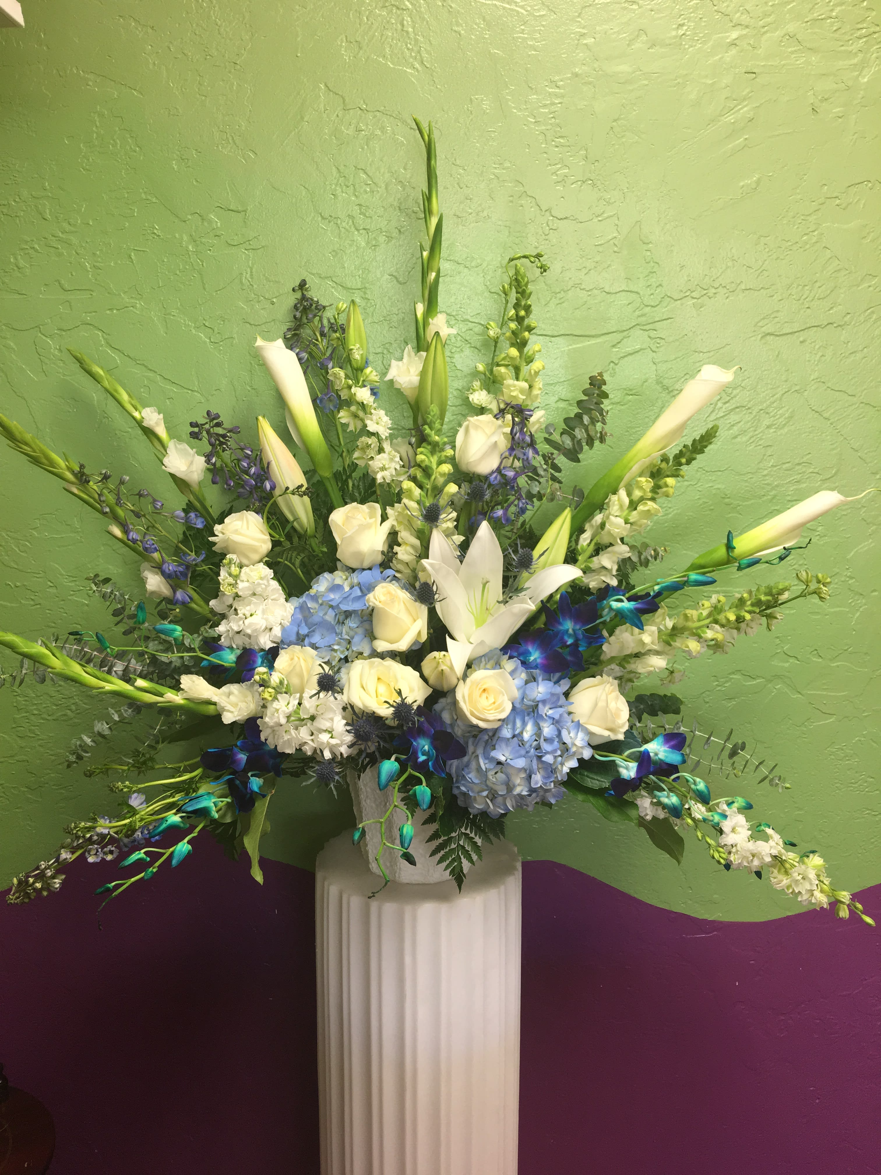 Tall floral arrangement with white lilies, roses, and blue hydrangeas in a white pedestal vase