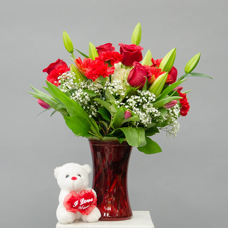Red bouquet in a glass vase with a small teddy bear holding a heart