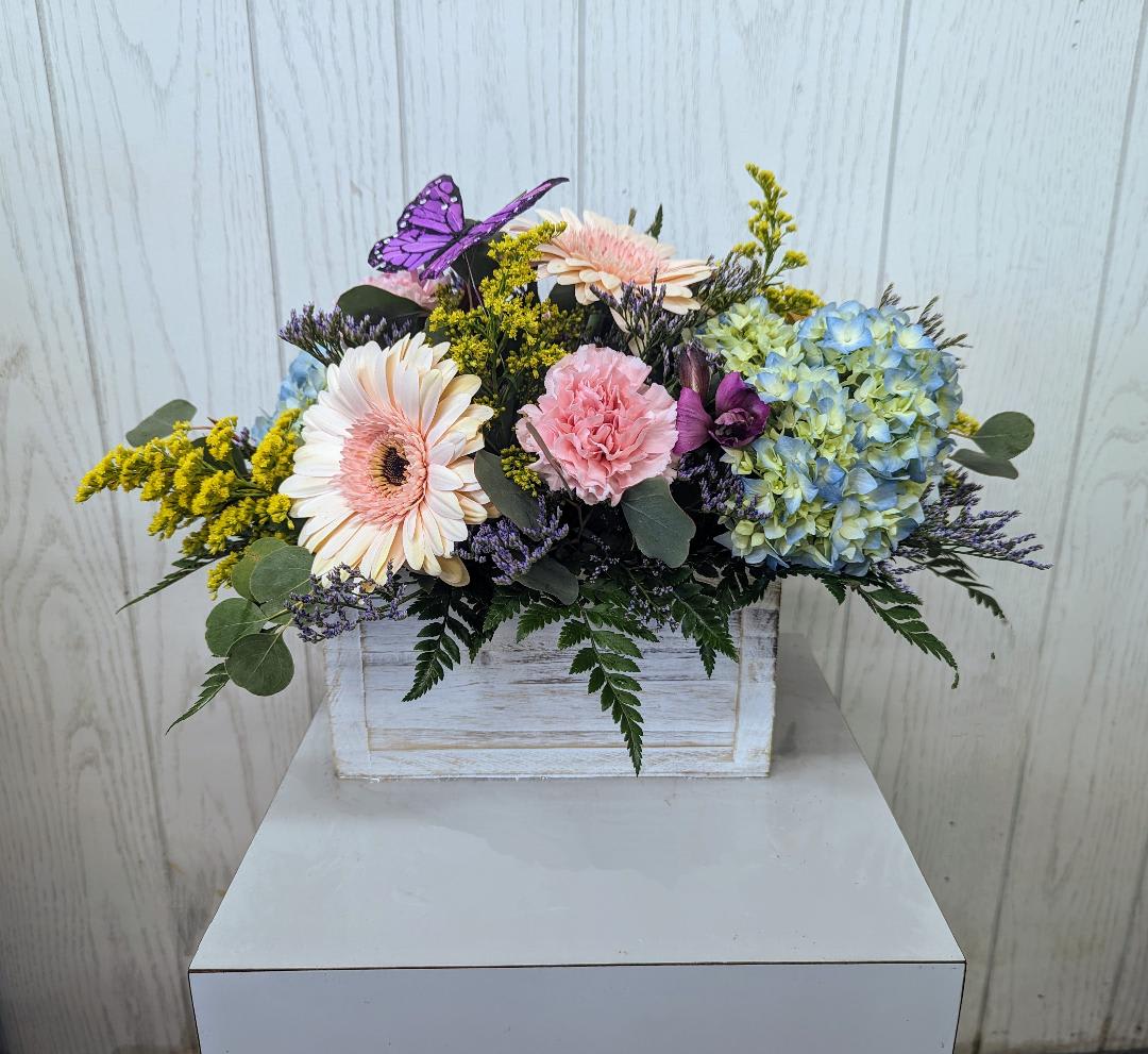 Mixed pastel flowers in a white wooden box with a purple butterfly accent.