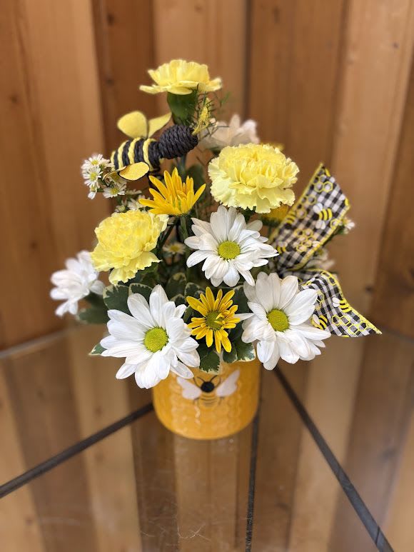 Bee Happy Bouquet - Bring a little buzz of happiness with our Bee Happy Bouquet! Arranged in an adorable bee-themed container, this charming design features sunny yellow carnations, bright white daisies, and pops of golden blooms, all nestled in fresh greenery. A cute bee accent and coordinating ribbon add a playful touch, making this arrangement perfect for spreading smiles, celebrating spring, or sending a sweet “thinking of you.” It’s the perfect pick-me-up that’s as cheerful as a sunny day!
