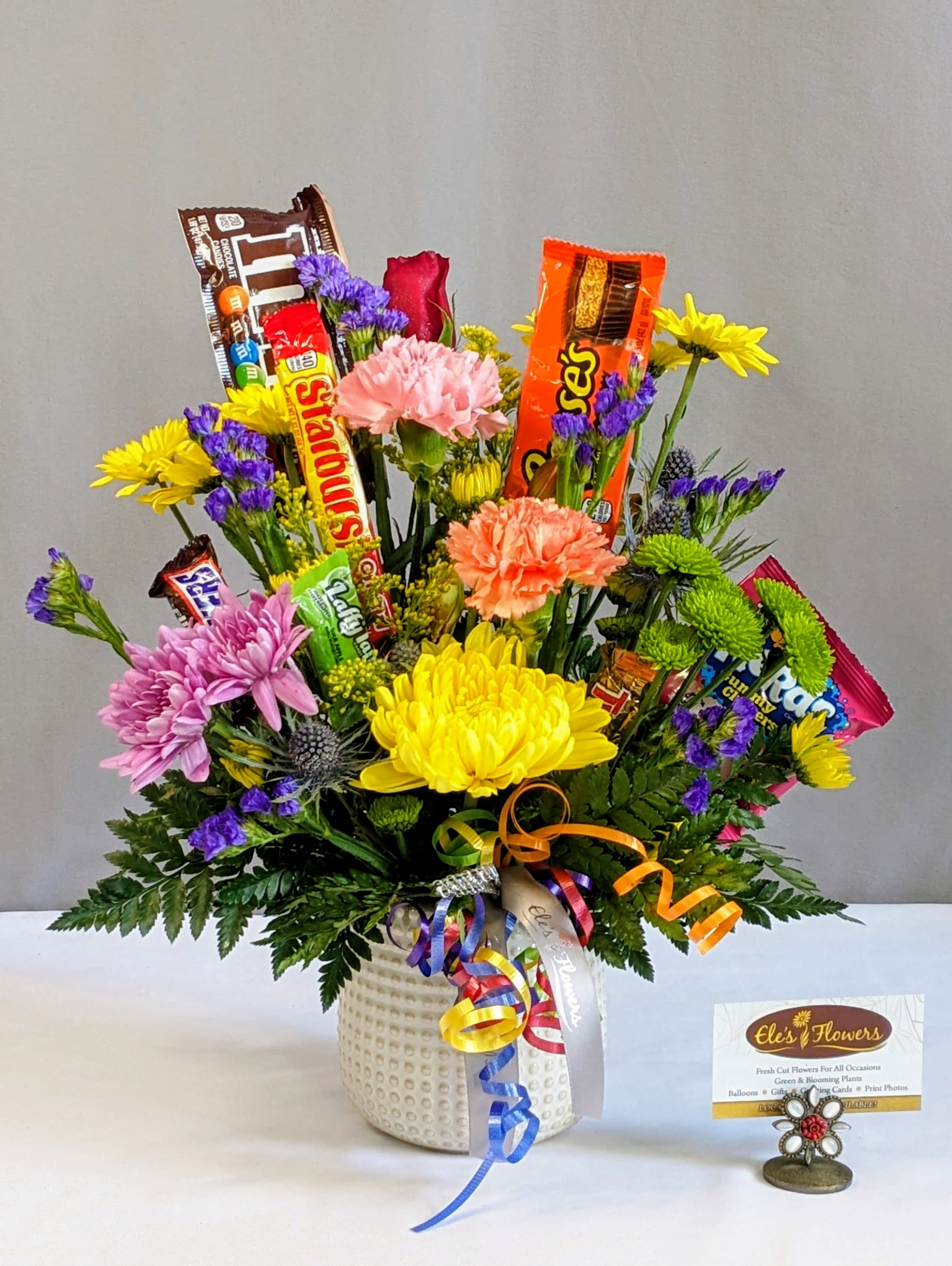 Confectionary Garden - This vibrant and joyful arrangement features a stunning mix of bright, rich, and bold primary color flowers that pop with energy and warmth. Adorned with an assortment of sweet candy bars and treats, it offers a delightful treat for both the eyes and the taste buds. Nestled in a container or vessel, the arrangement is complemented by fresh, lush foliage, adding a natural, verdant touch. Perfect for celebrations such as birthdays, congratulations, get well wishes, or simply as a cheerful "just because" gesture. Please note that blooms and sweet treats may vary based on availability, ensuring a unique and fresh touch in every arrangement.  Approximate Dimensions: 16"H x 13"W