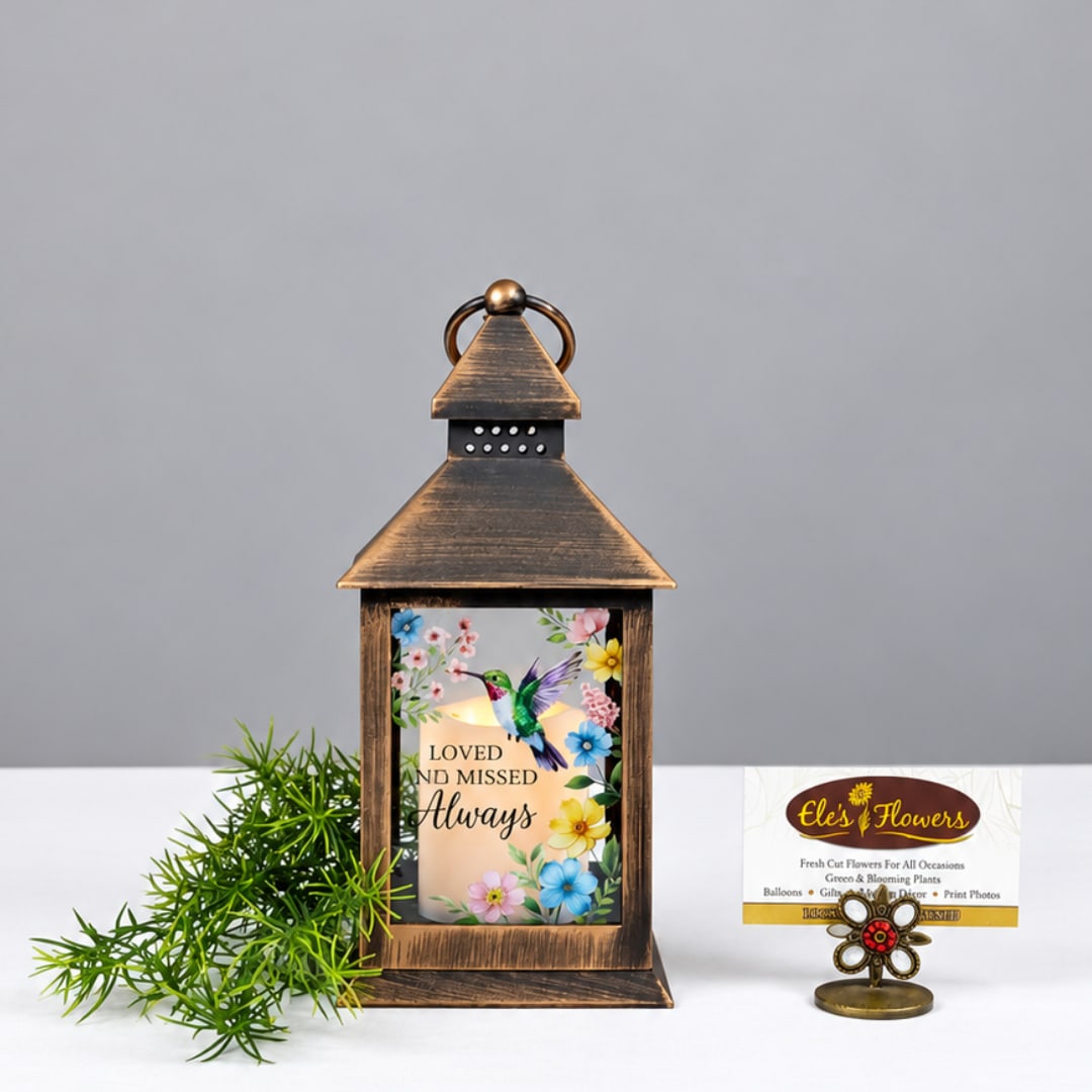 Bronze lantern with a memorial candle and floral print, beside greenery and a small card holder