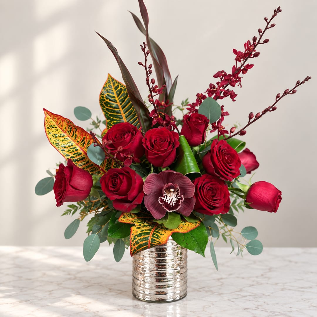 Red roses arranged in a silver vase with burgundy orchids and patterned leaves