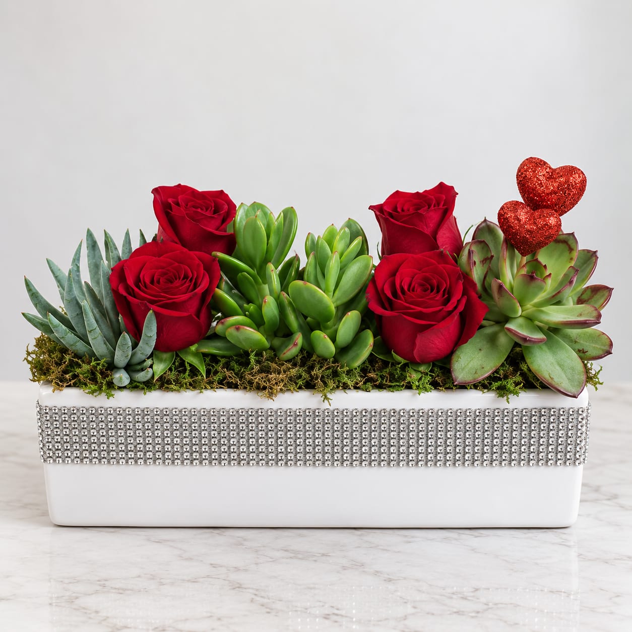 Red roses and succulents arranged in a white rectangular planter with glitter hearts