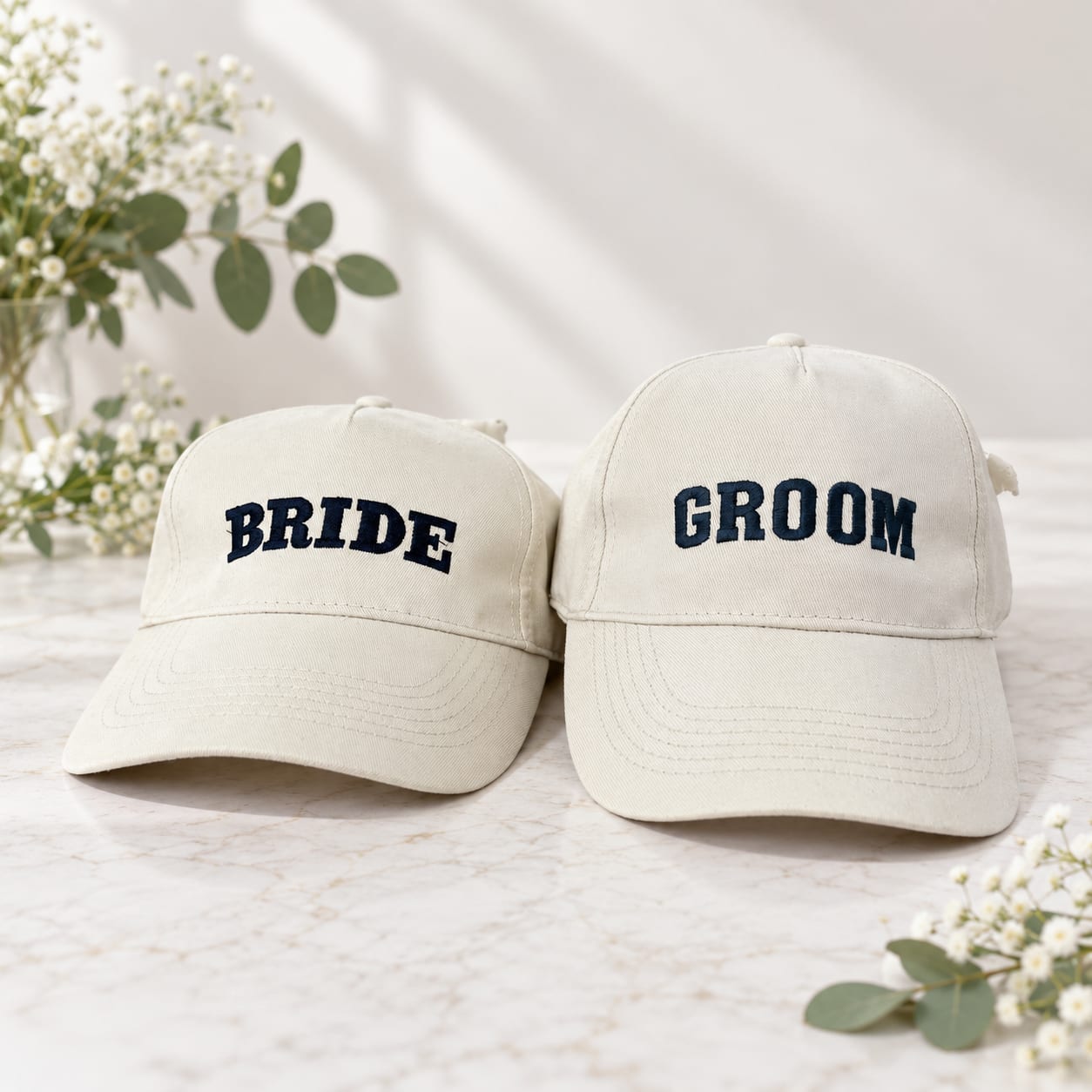 Two cream caps embroidered Bride and Groom on a marble surface