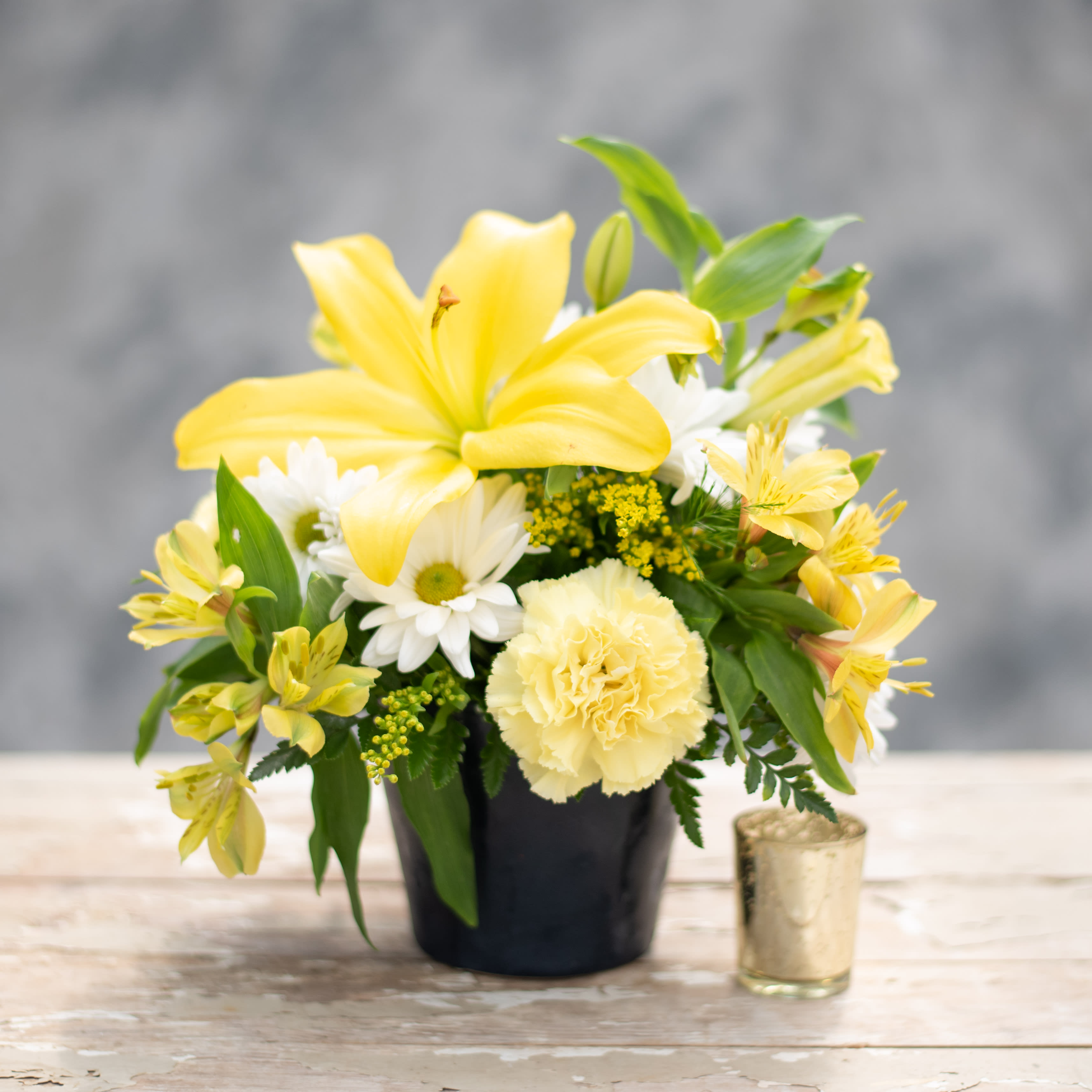 Yellow lilies and white daisies in a black pot with a small candle beside it