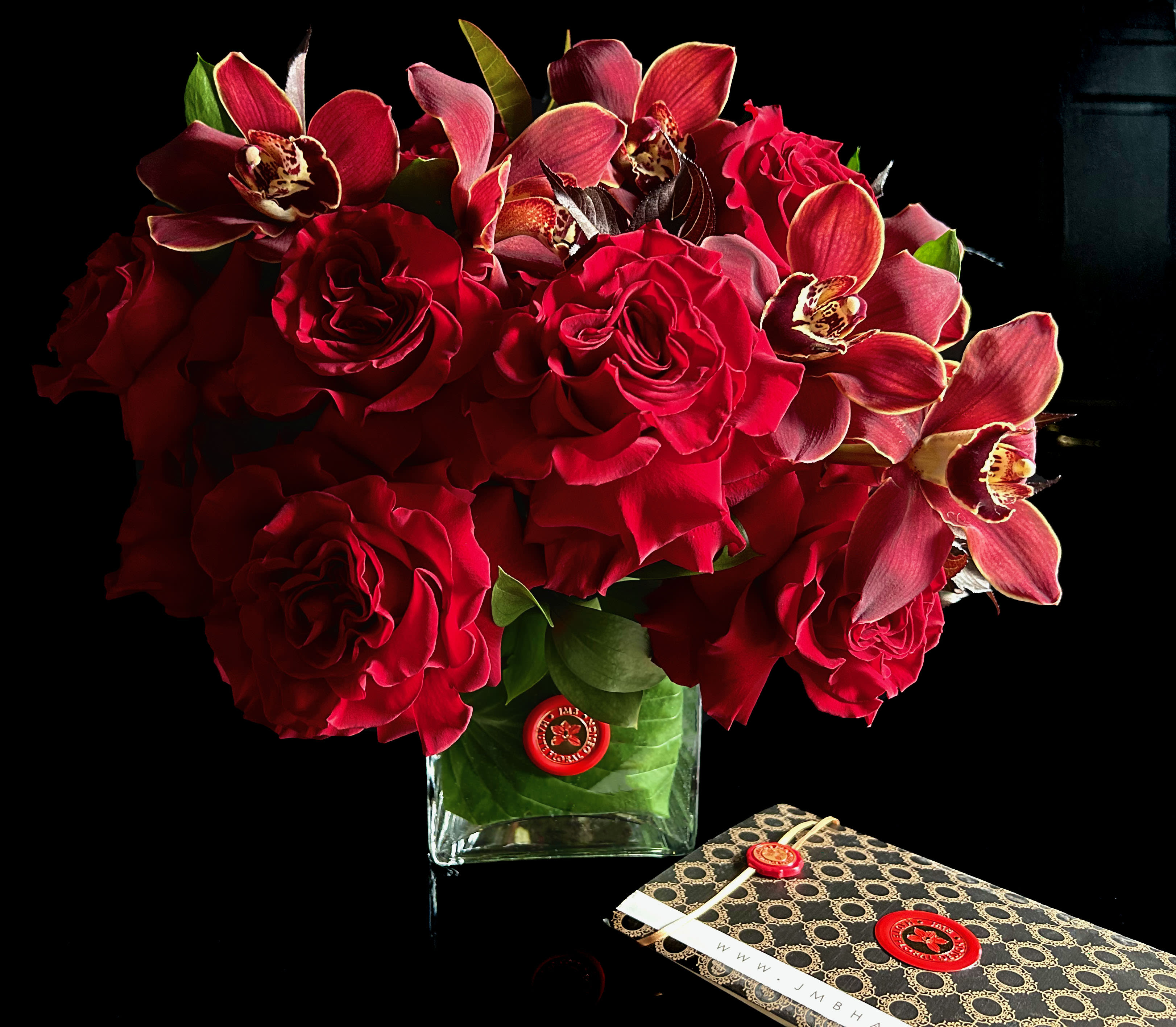 Red roses and orchids arranged in a clear square vase beside a patterned gift box