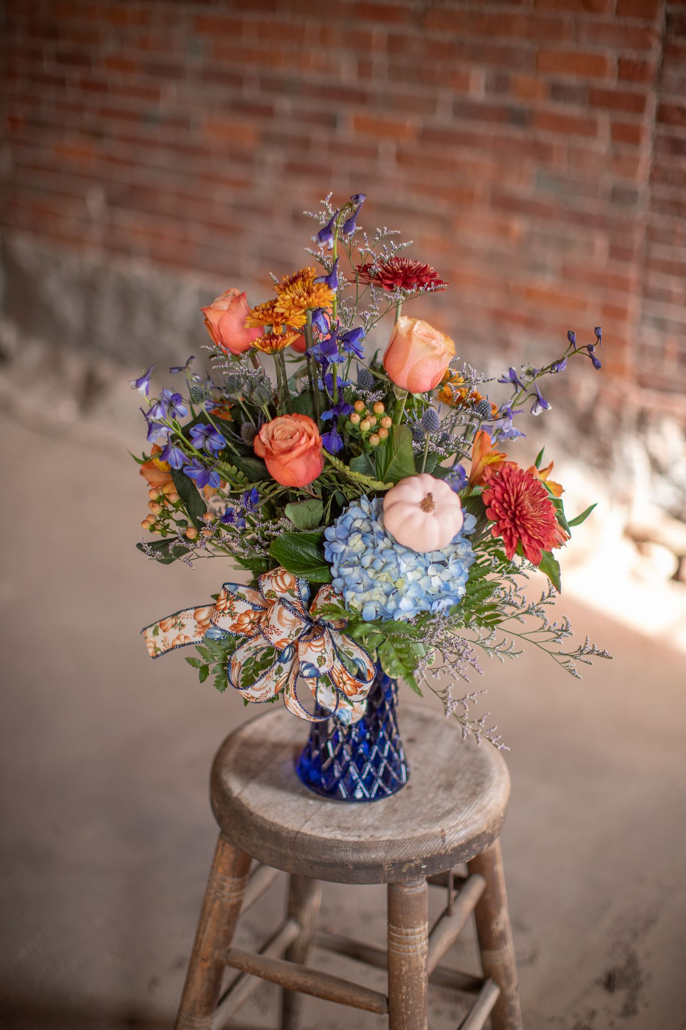 Country Pumpkins II - An upgraded version of the season's favorite, "Country Pumpkins," is ready for this fall. Bicolor terracotta-peach roses with butterscotch buttons, blue hydrangea, thistle and more is complete with a festive linen bow printed with matching pumpkin print. All these things and more are artfully arranged in a texture cobalt blue gathering style vase.  