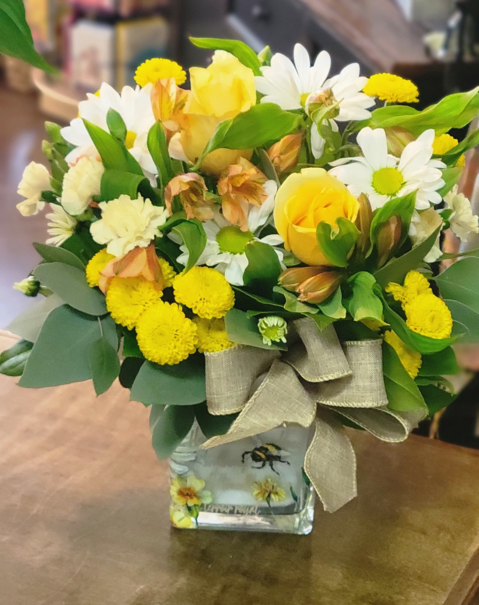 Yellow and white mixed flower arrangement with roses and daisies in a bee-themed glass cube vase with a burlap bow
