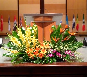Large tropical stage arrangement with yellow spikes, orange lilies, and birds of paradise before a podium