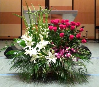 Large stage arrangement of white lilies, hot pink roses, and purple flowers with tall green spikes in a low container.