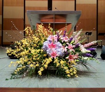 Large stage floral arrangement with pink lilies, lavender hydrangeas, yellow orchids, and tall accent stems