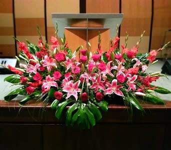 Large altar spray of pink lilies and red roses with bold tropical leaves on a wooden podium.