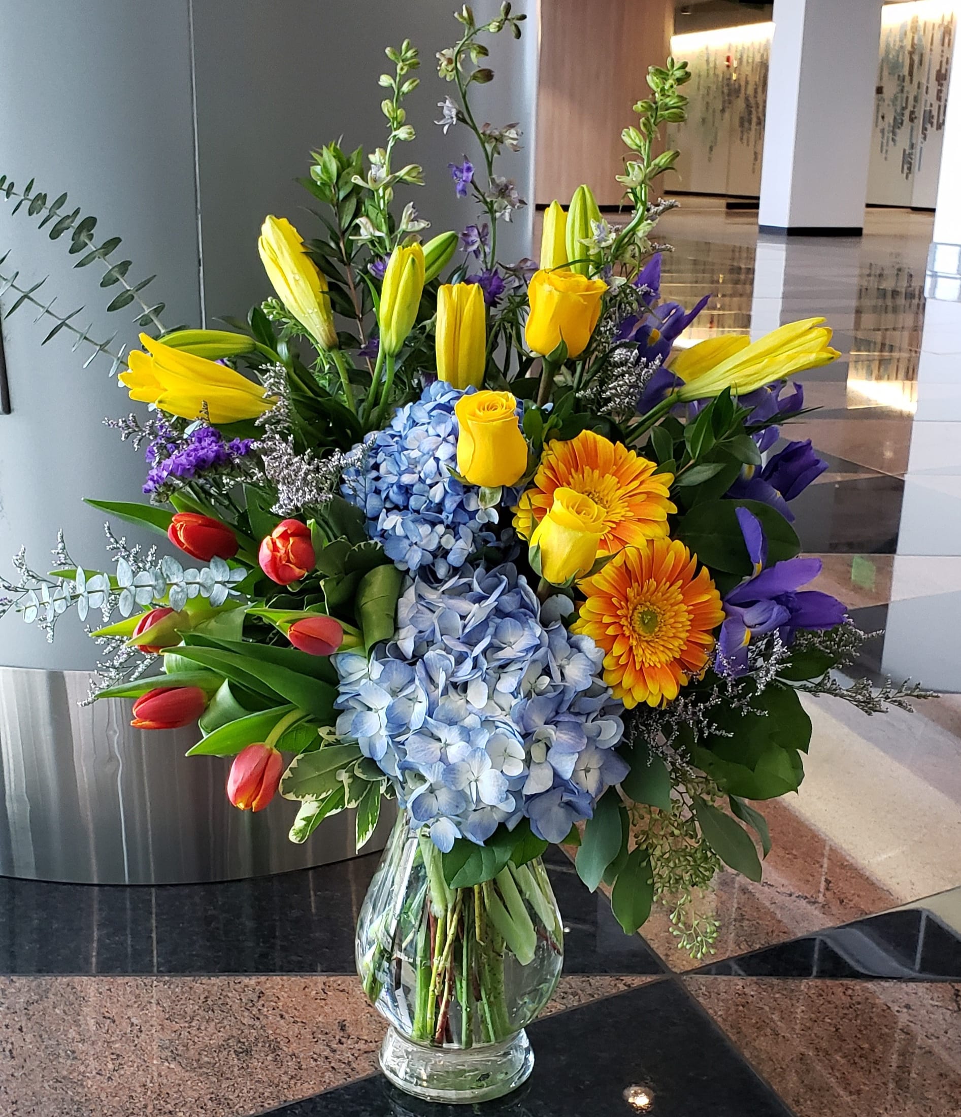 Colorful mixed bouquet in a clear glass vase with tulips, roses, hydrangeas, and gerbera daisies