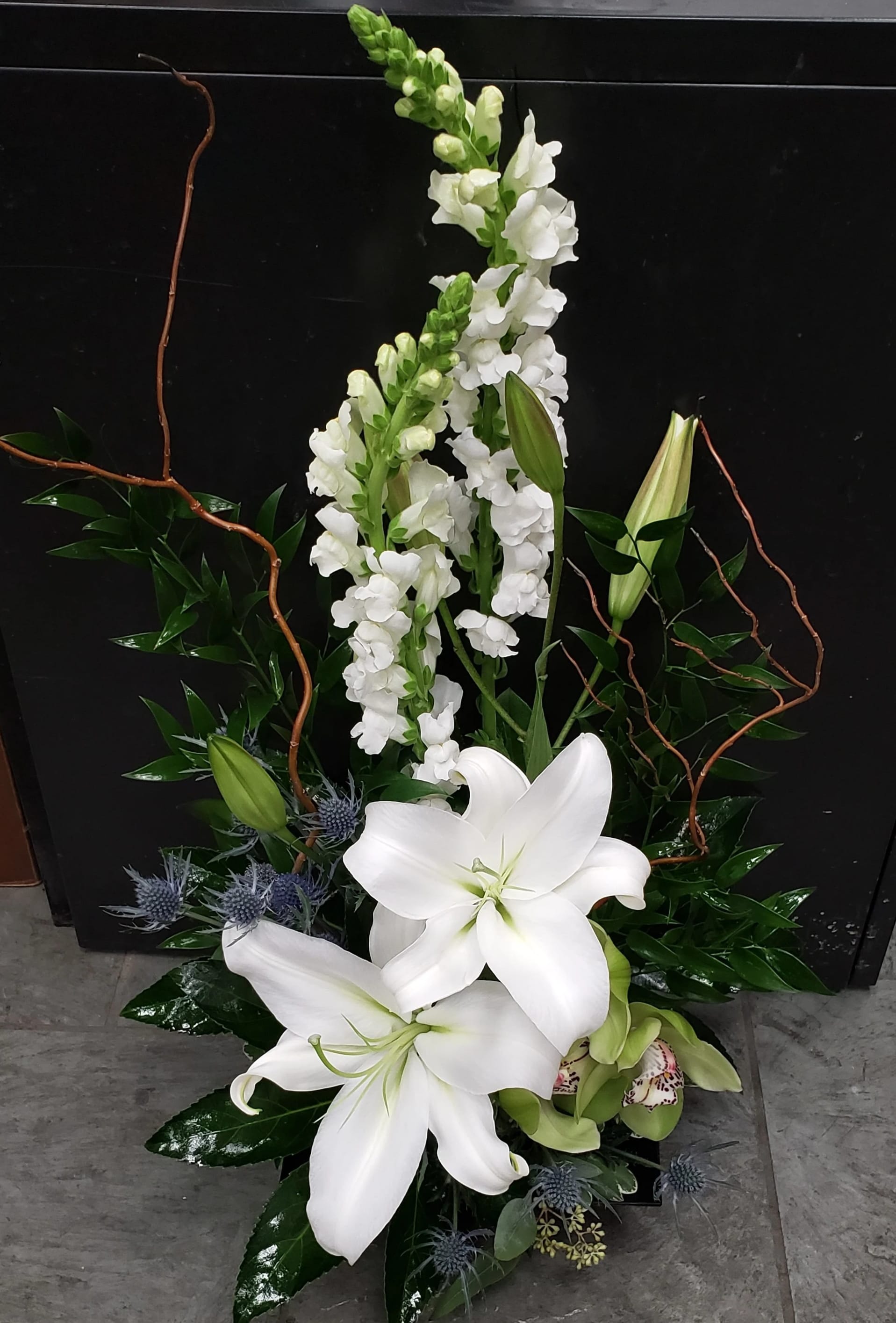 Tall arrangement of white lilies and snapdragons with blue thistle in a dark container