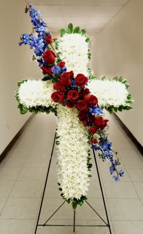 Tall white floral cross with red roses and blue flowers on a metal stand