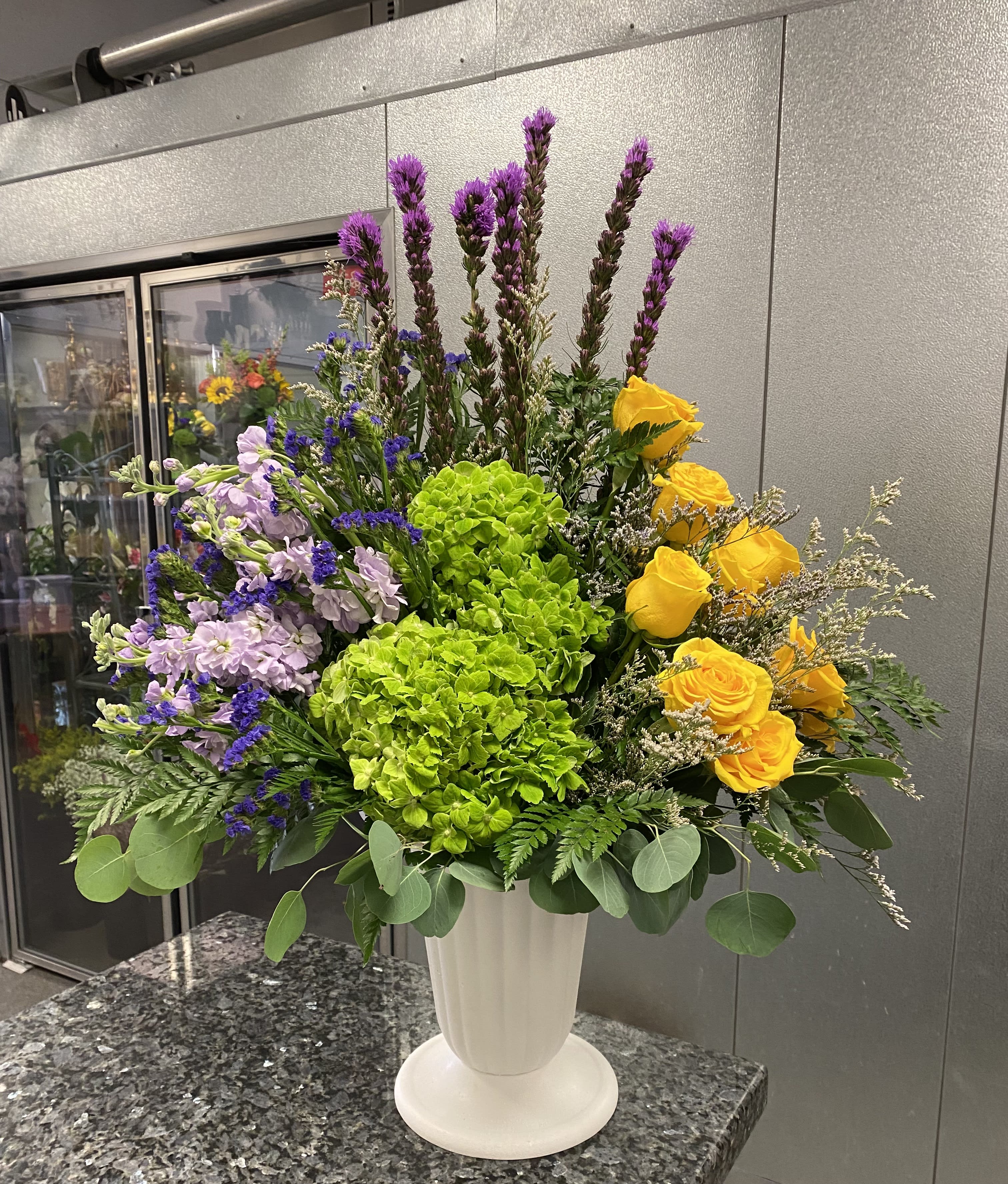 Tall floral arrangement with yellow roses, green hydrangeas, and purple flowers in a white vase