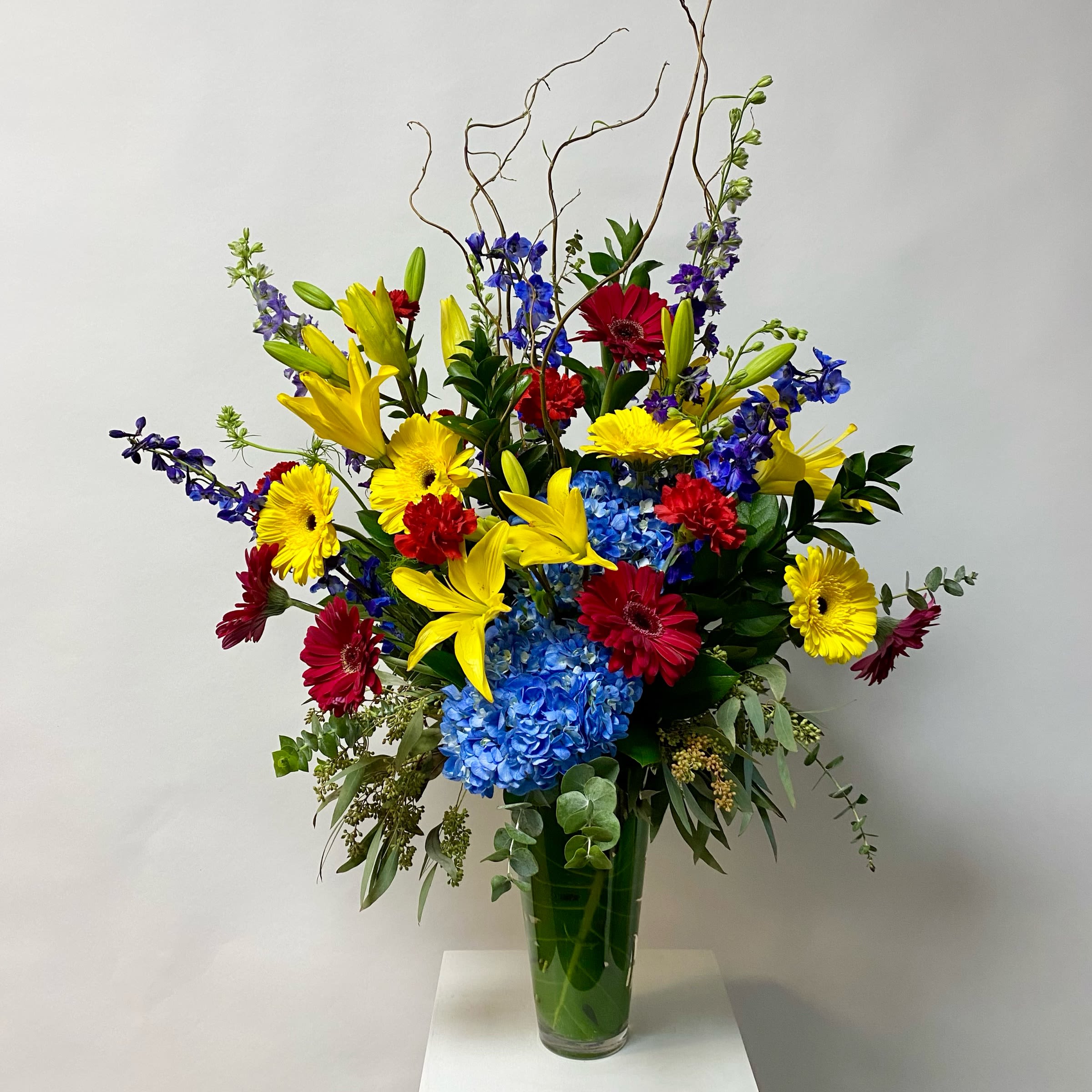 Tall mixed bouquet in a glass vase with yellow lilies, gerberas, blue hydrangea, and red blooms