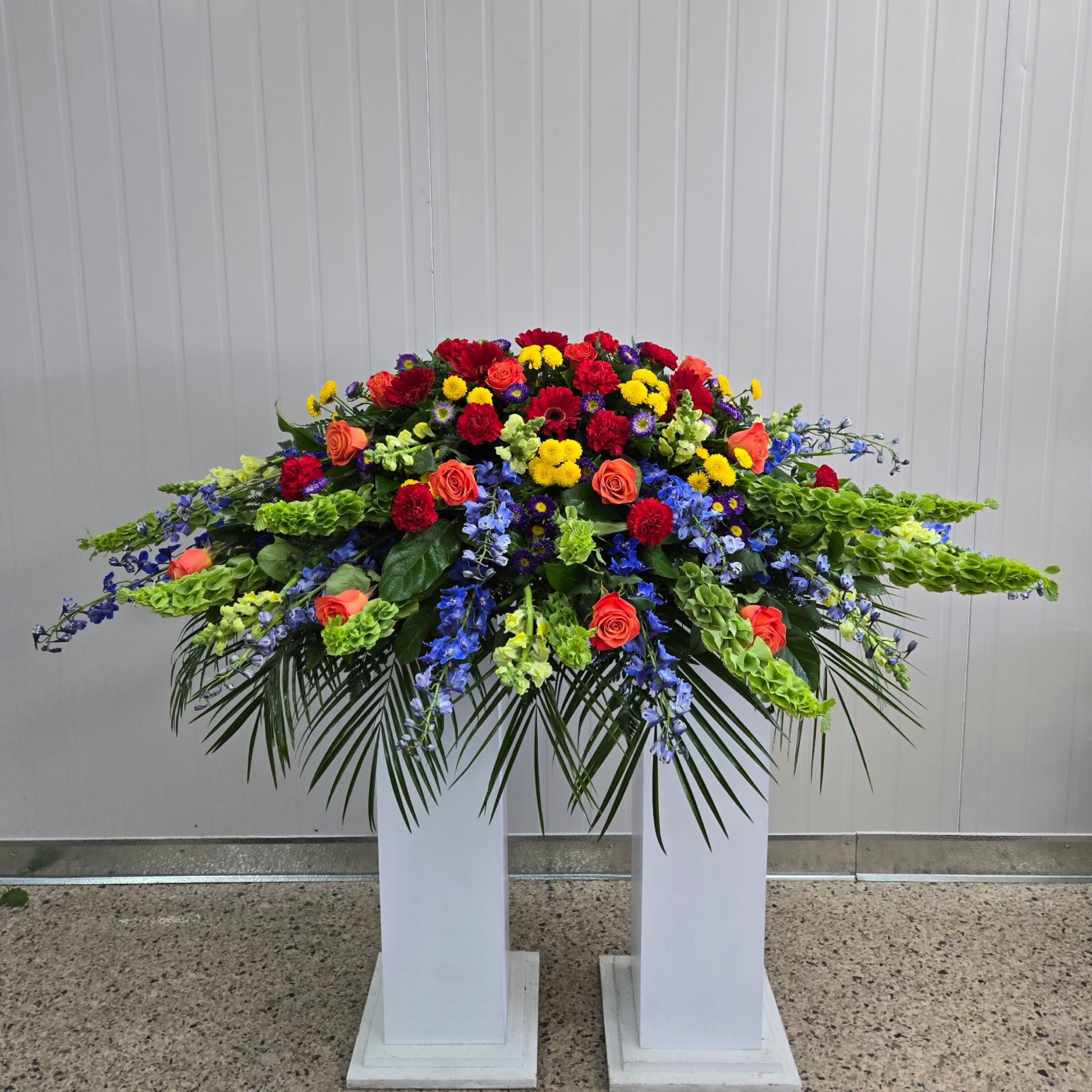 Vibrance Casket Spray - This Casket Spray Includes Red Gerbera Daisy, Bells of Ireland, Blue Delphinium, Purple Matsumoto, yellow Snapdragon, Red Carnations, and Orange Roses. Or as Similar as Possible. 