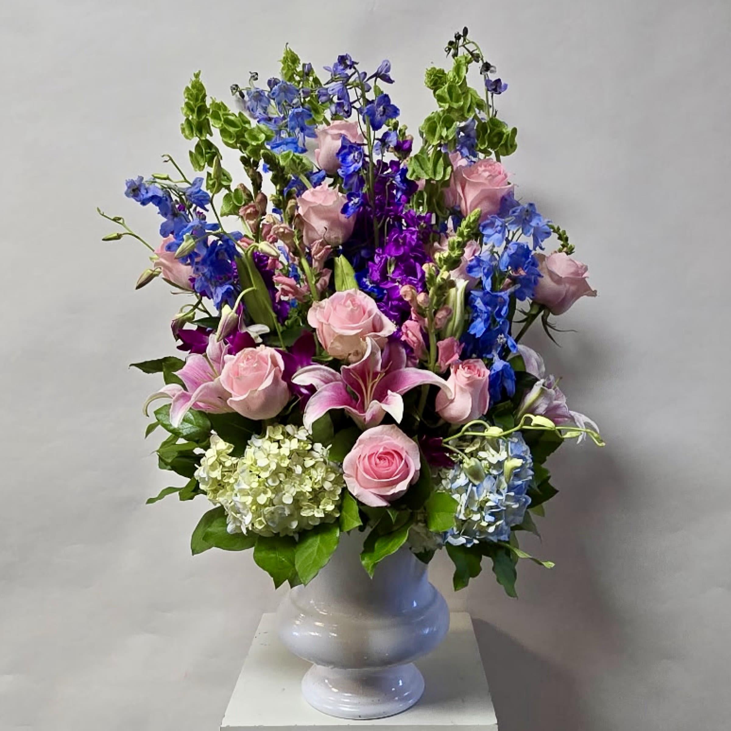 Loving Memories Arrangement - This Arrangement Includes Bells of Ireland, Purple Dendrobium Orchids, Light Blue Delphinium, Blue Hydrangea, Purple Stock, Pink Lily, Light Pink Snapdragon and Light Pink Rose in a Mortuary approves Plastic Container. Or as Similar as Possible. 